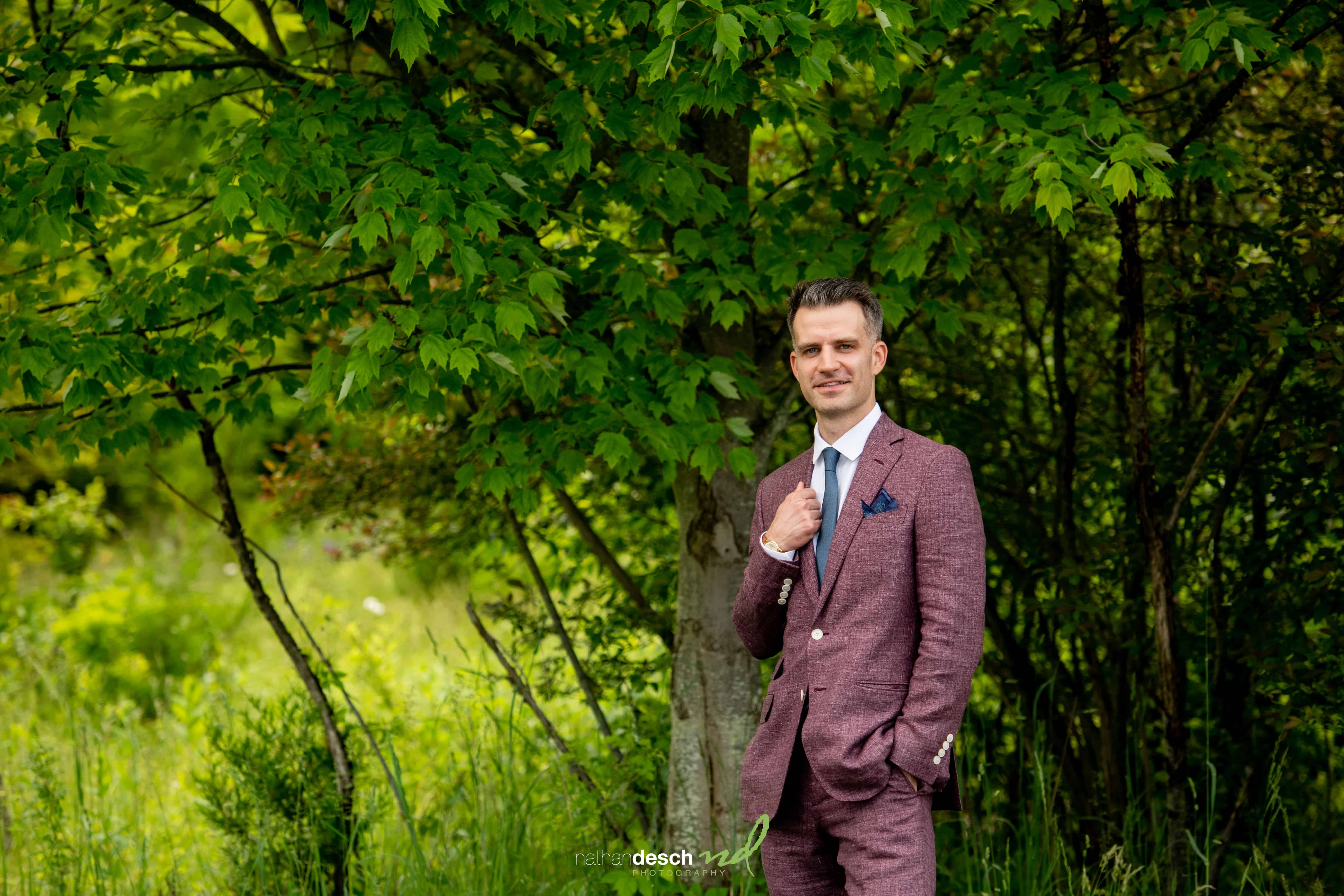 Groom in maroon suit smiling outdoors among green trees and grass, holding tie on Will and Laura’s wedding day.