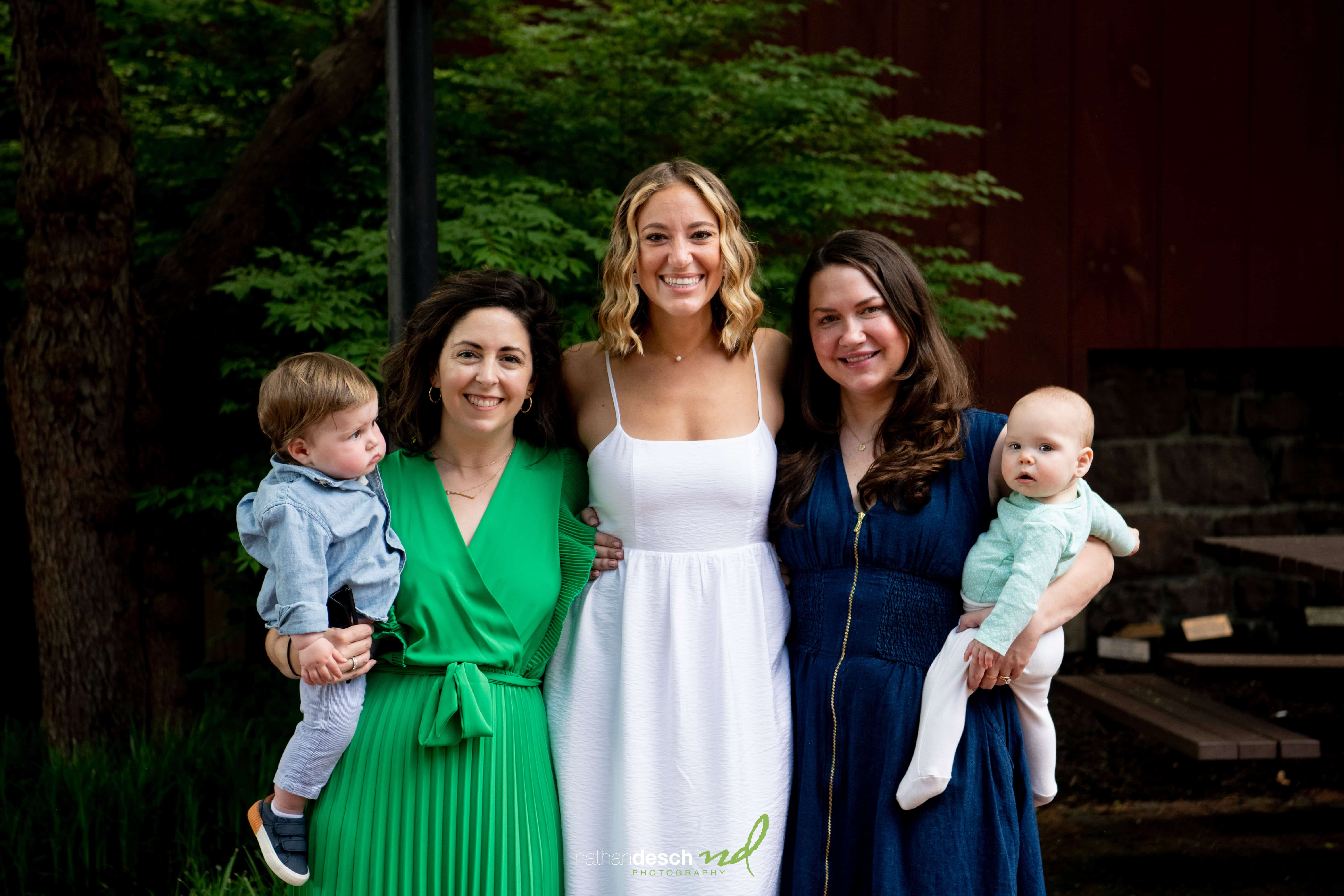 Bride in white dress smiles with bridesmaids in green and blue dresses, each holding babies, outdoors with lush greenery.