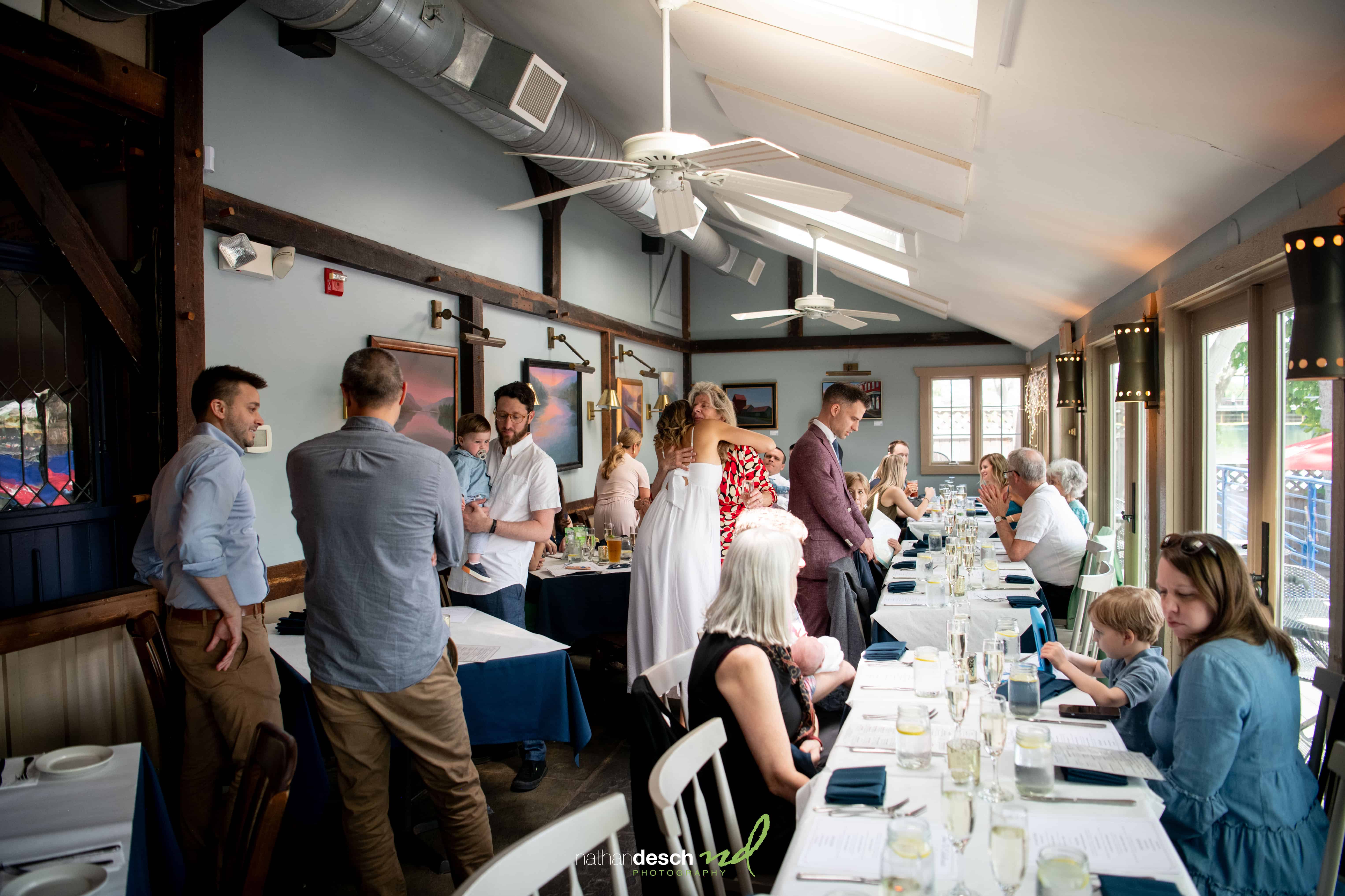 Laura and Will celebrate with guests around a sunlit dining table in an airy restaurant during their wedding reception.
