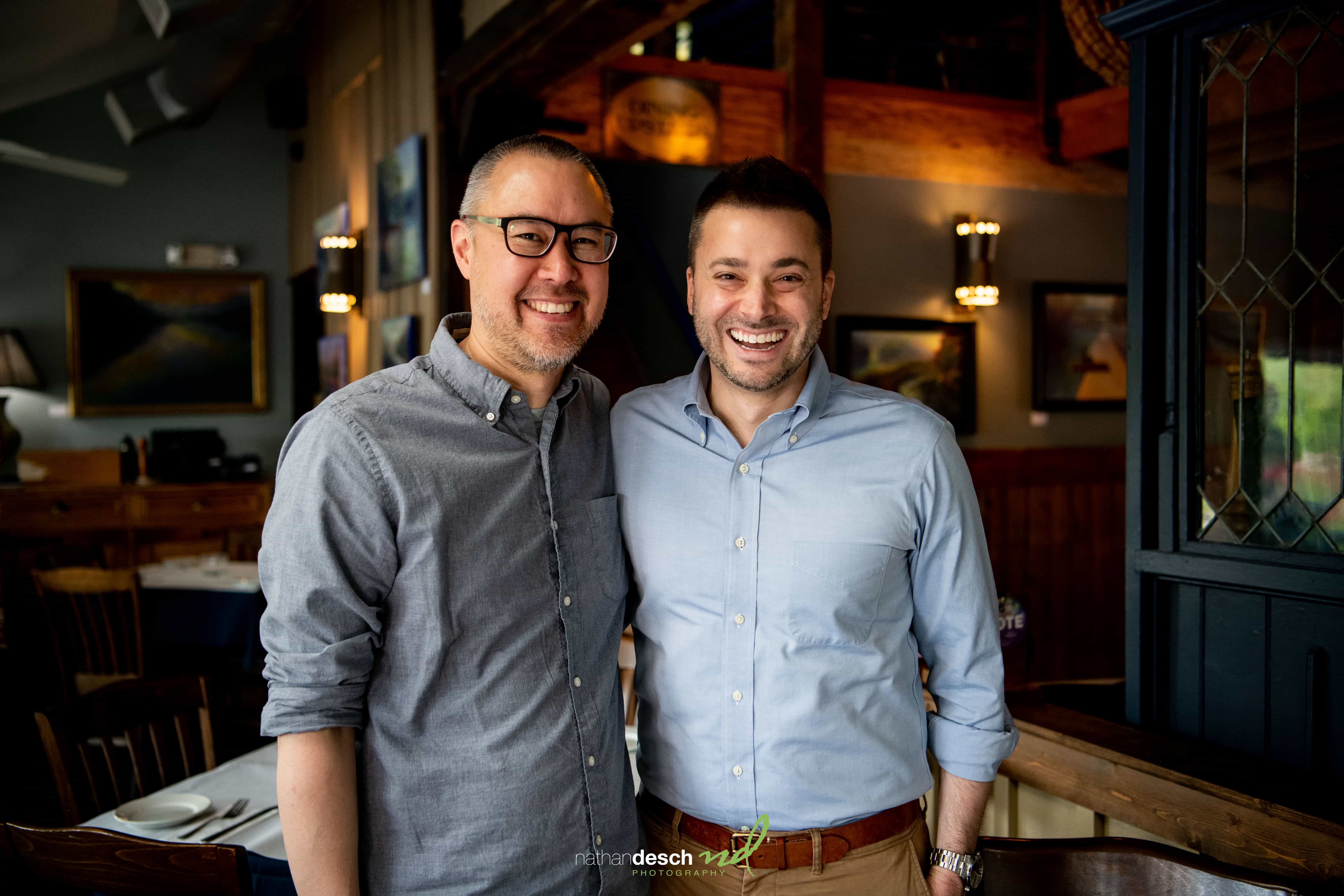 Two men in button-up shirts and glasses smile together inside a cozy restaurant wedding venue.