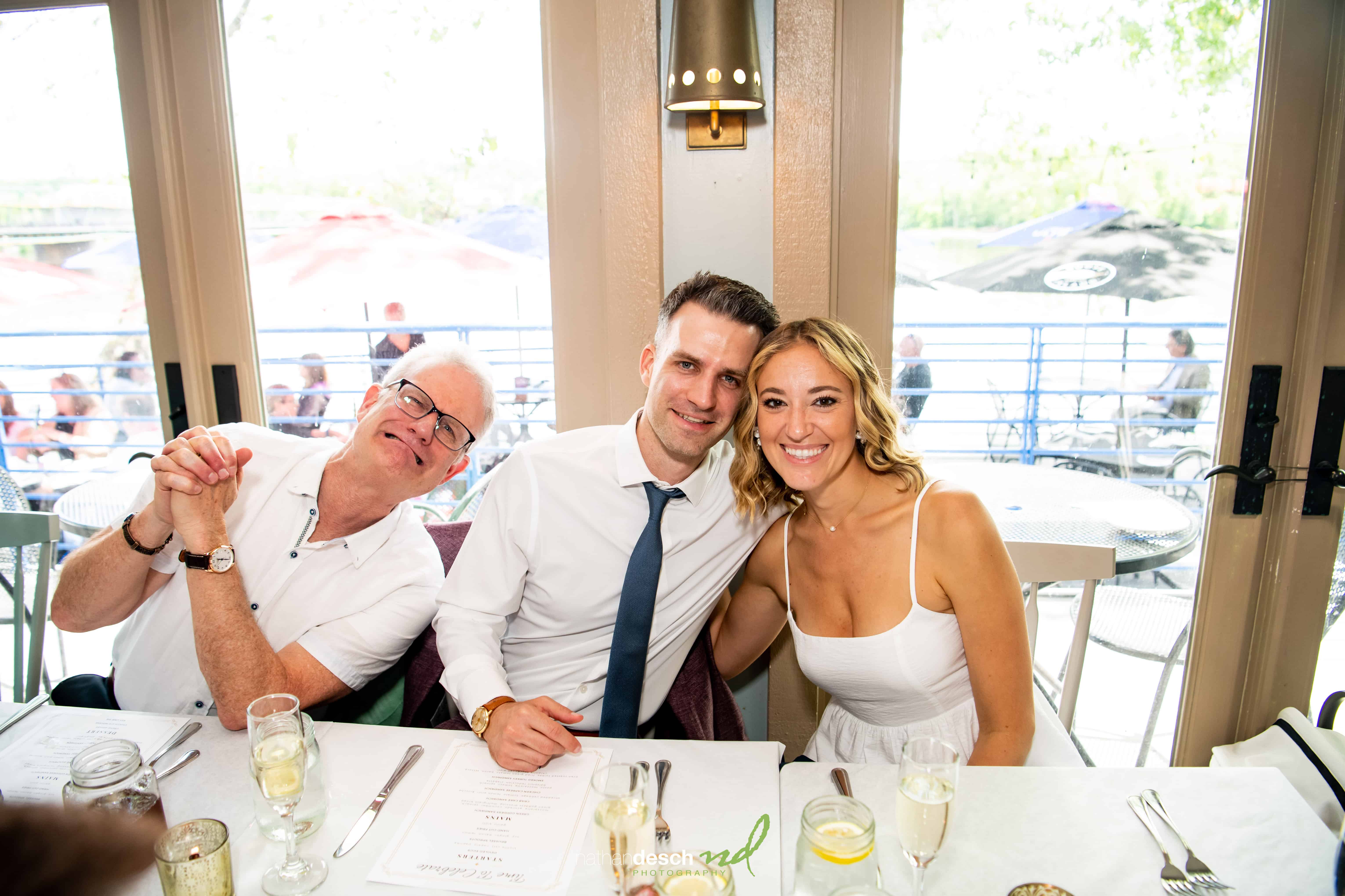Bride and groom smiling at restaurant table with a friend during wedding reception, candid joyful moment.