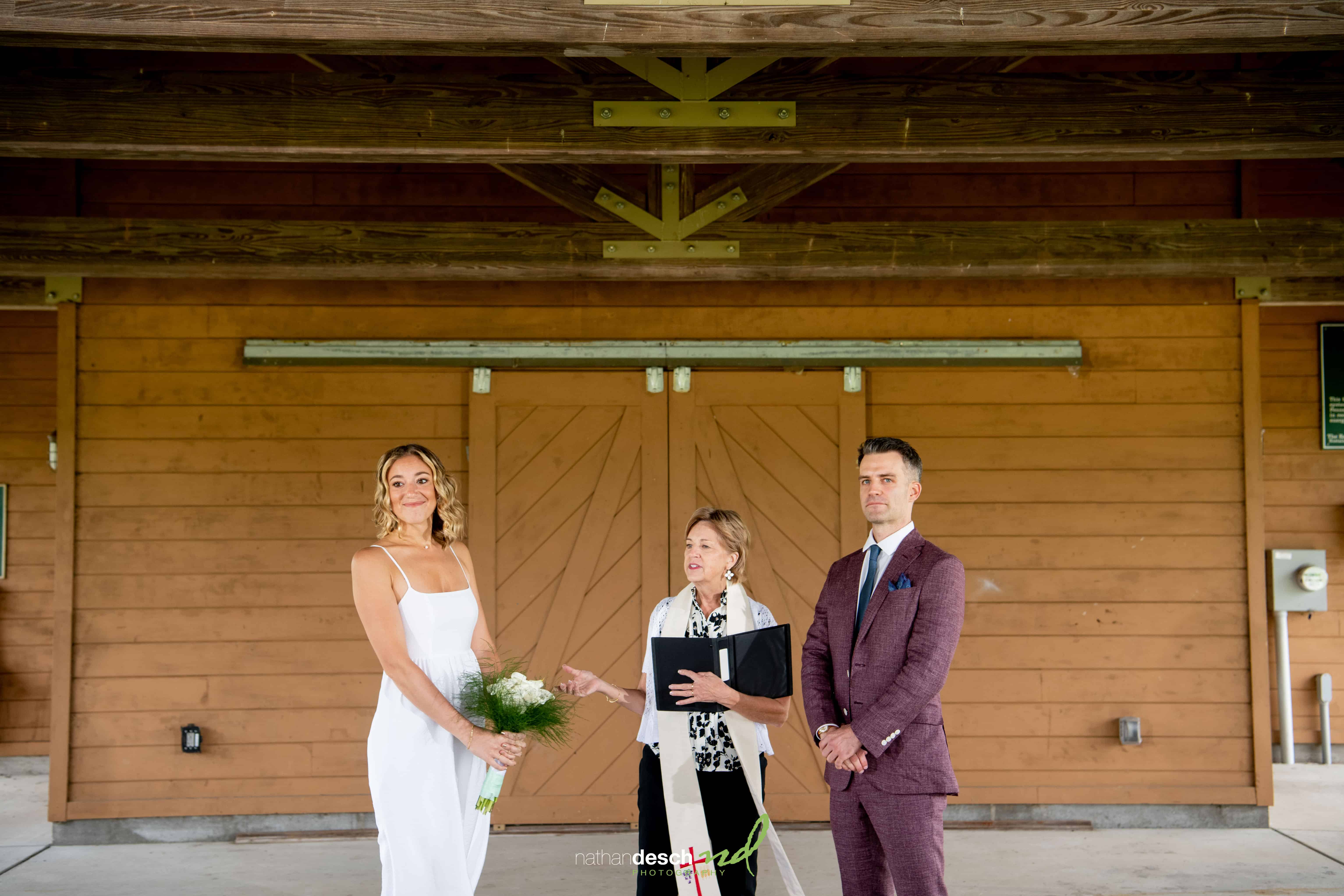 Bride in white dress with bouquet smiles beside groom in maroon suit and officiant outside rustic wooden wedding venue.