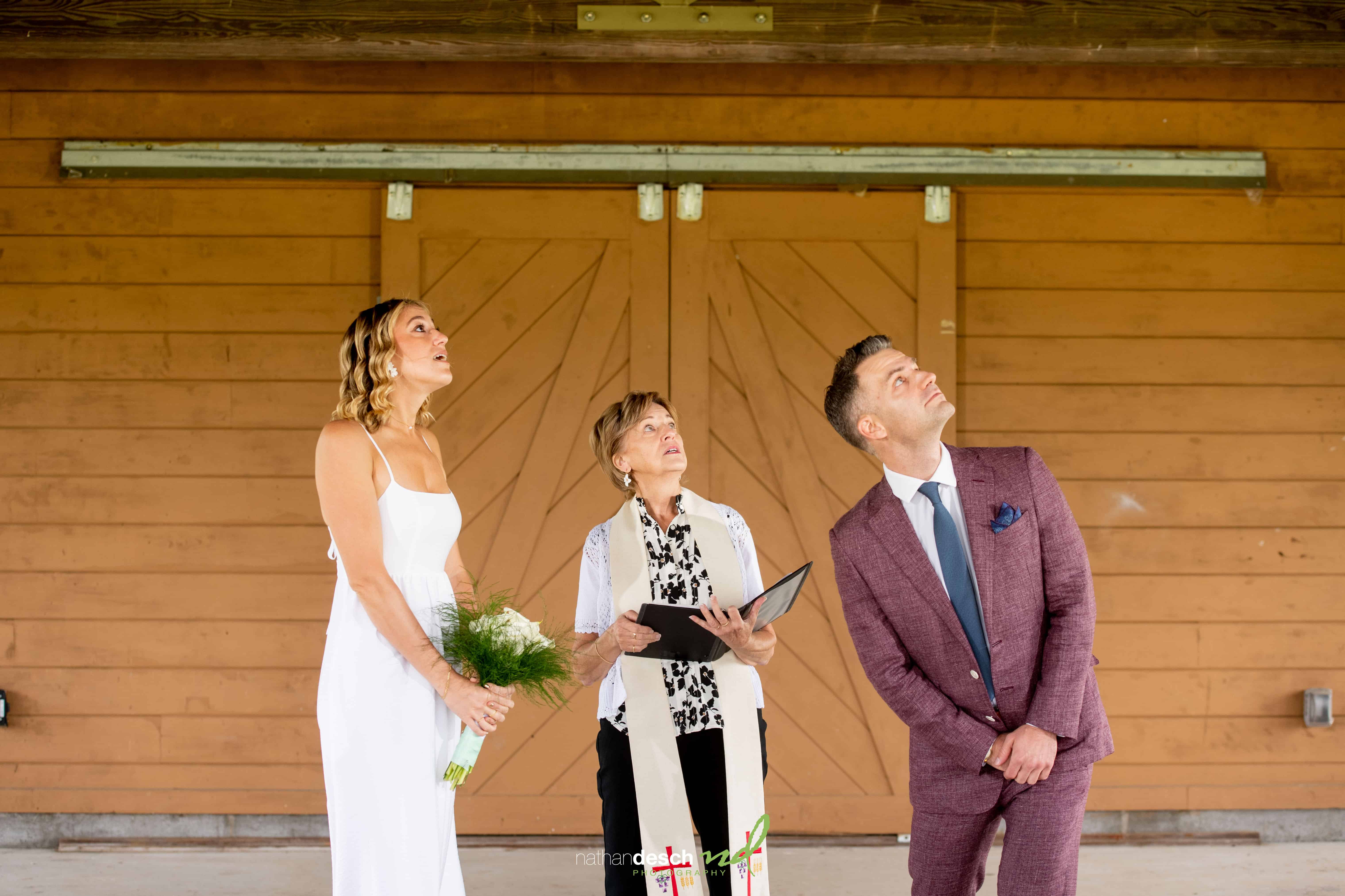 Bride and groom with officiant in front of wooden doors, bride in white dress holding bouquet, groom in maroon suit.