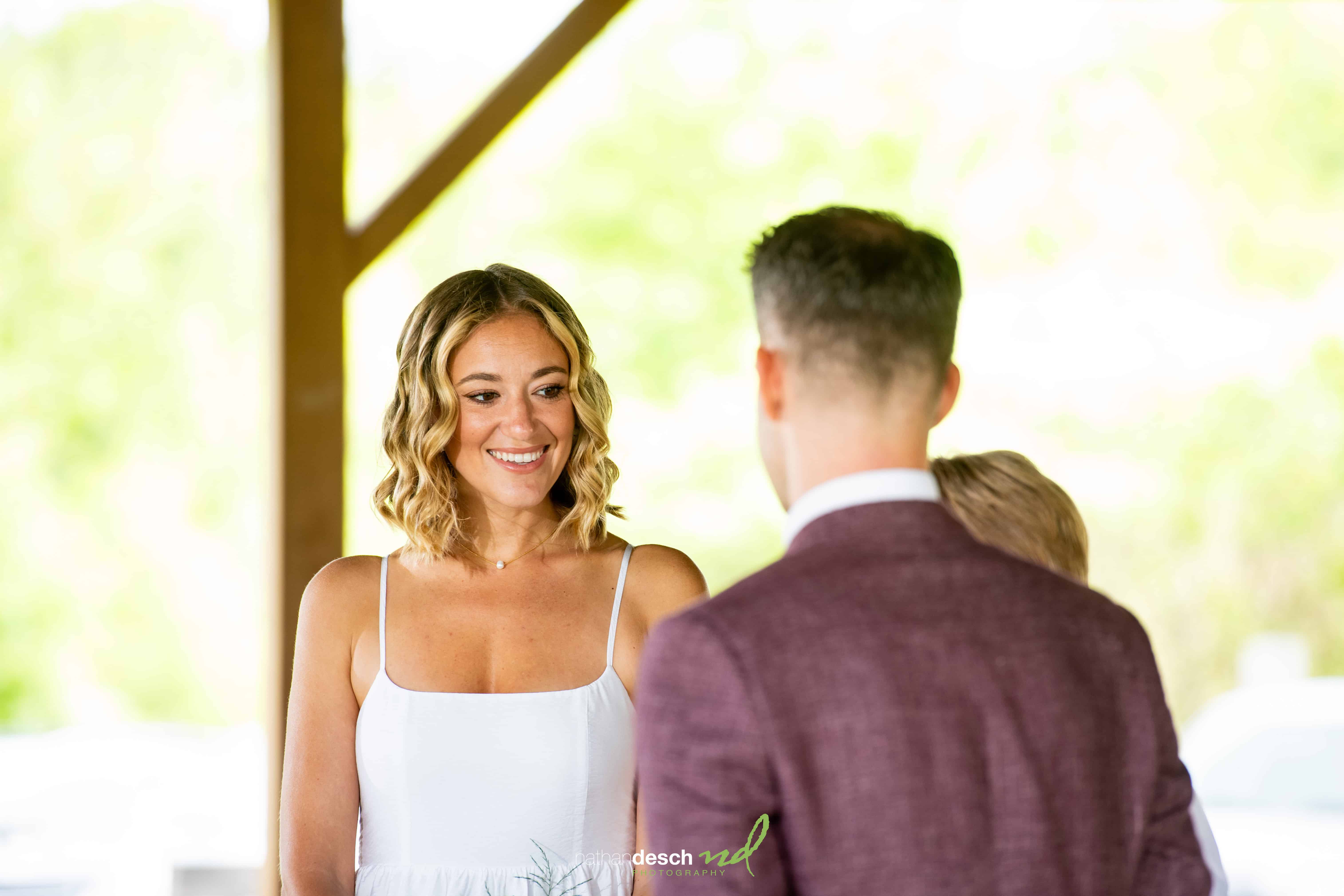 Bride in white dress smiles at groom in maroon jacket outdoors on a bright, sunny day with a blurred background.