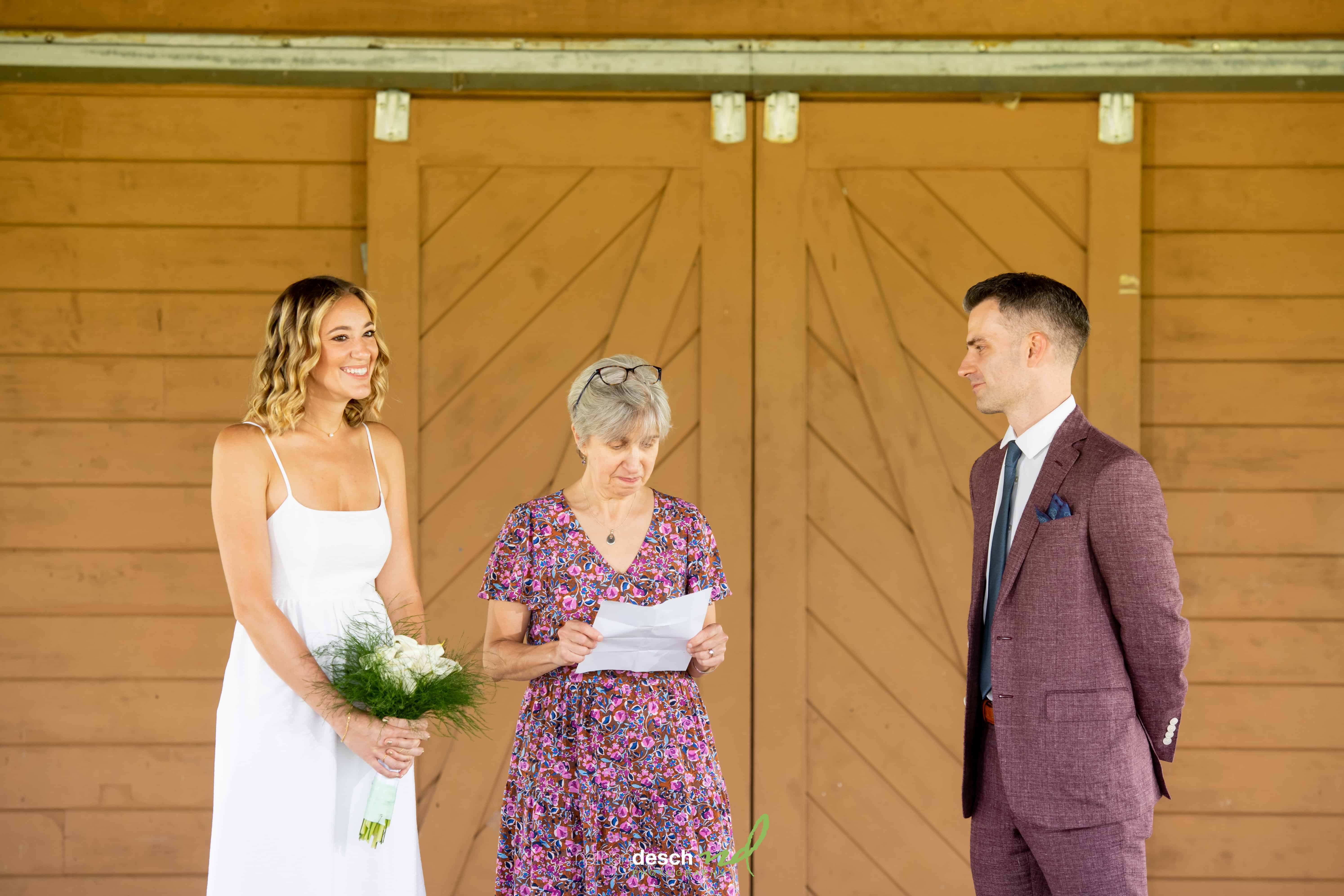 Bride in white dress with bouquet stands with smiling officiant and groom in maroon suit before rustic wooden doors on wedding day.