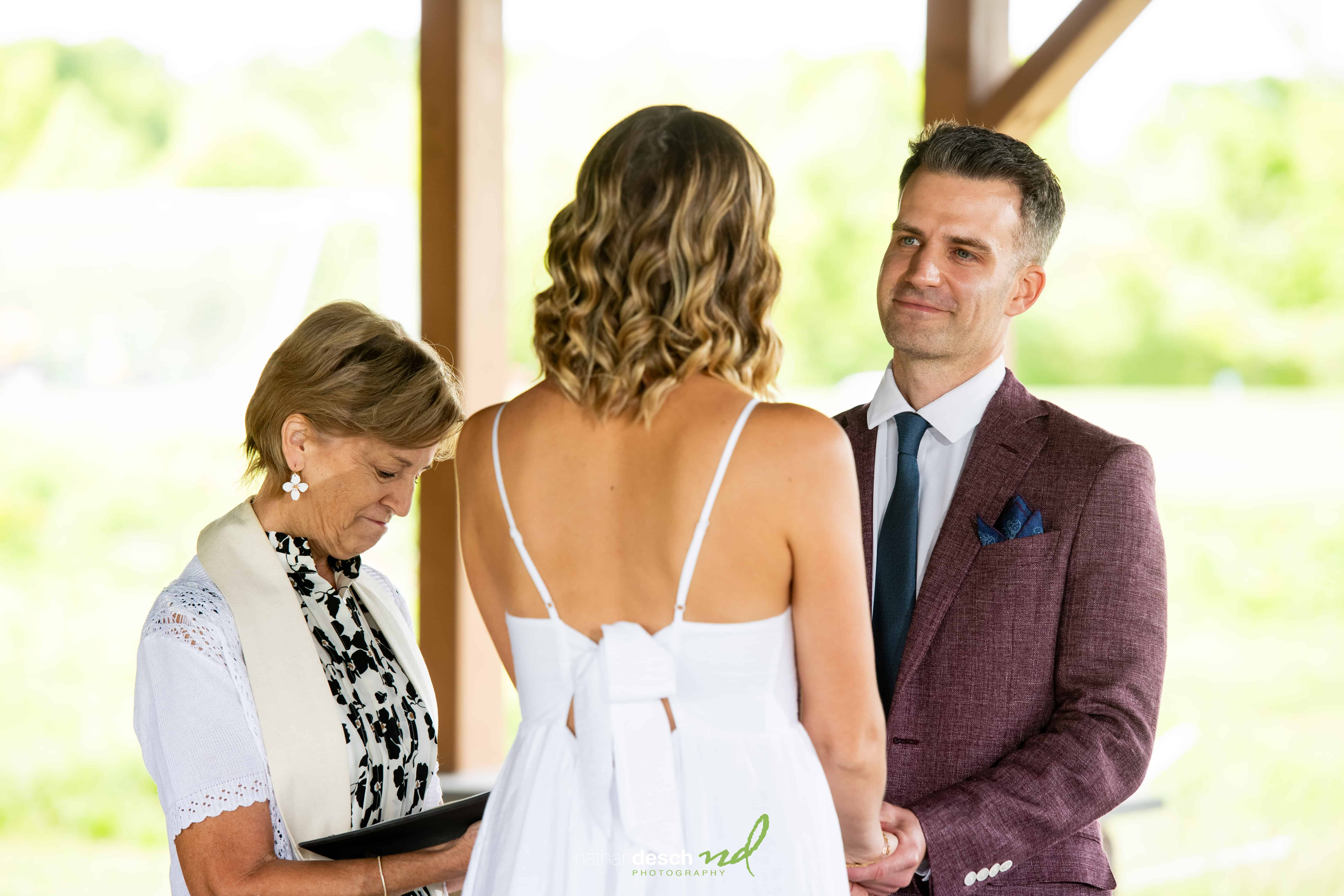Bride in white dress and groom in maroon suit hold hands during outdoor wedding ceremony with green, natural backdrop.
