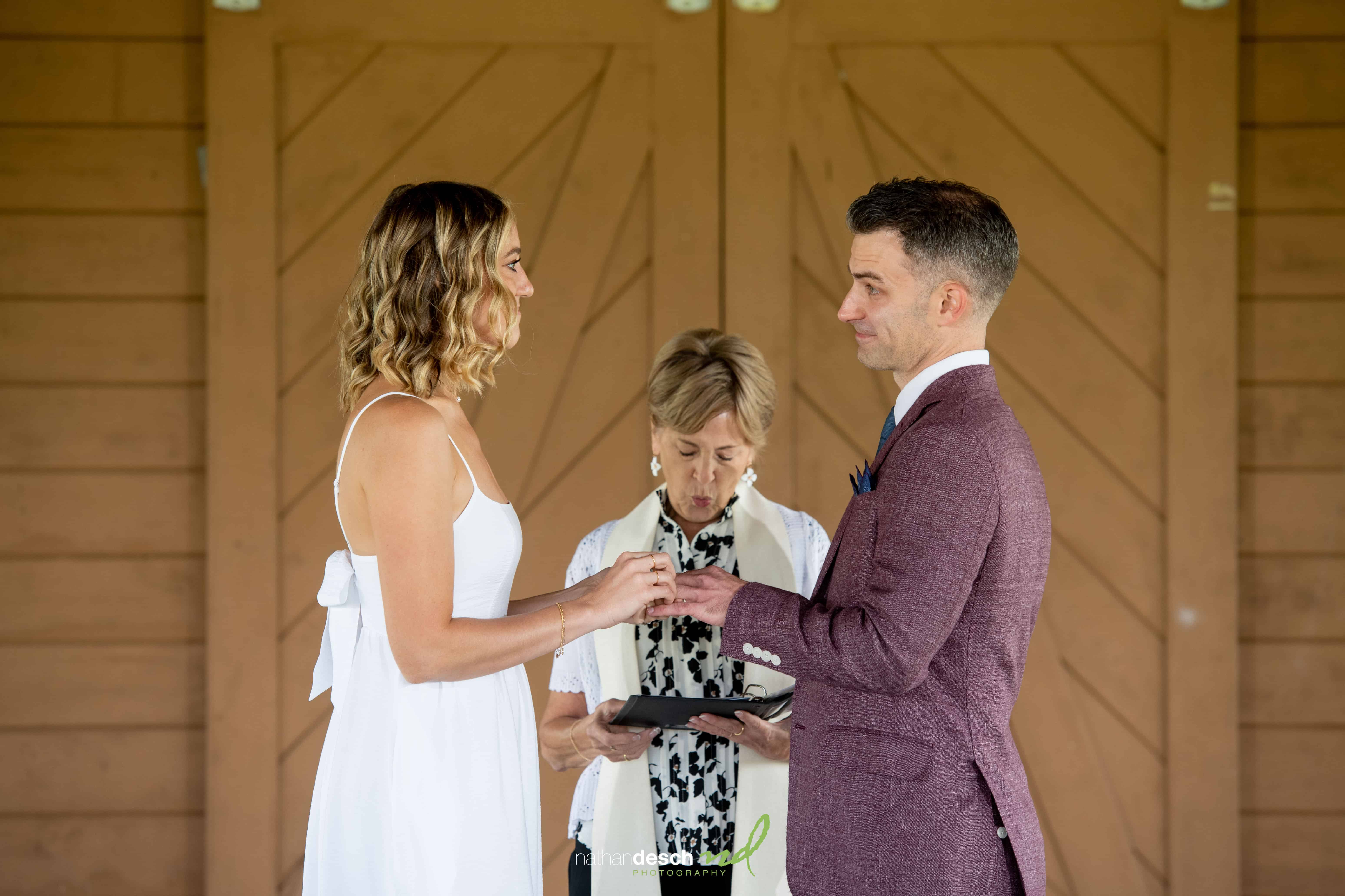 Bride and groom exchange rings before officiant during indoor wedding ceremony with rustic wooden doors in the background.
