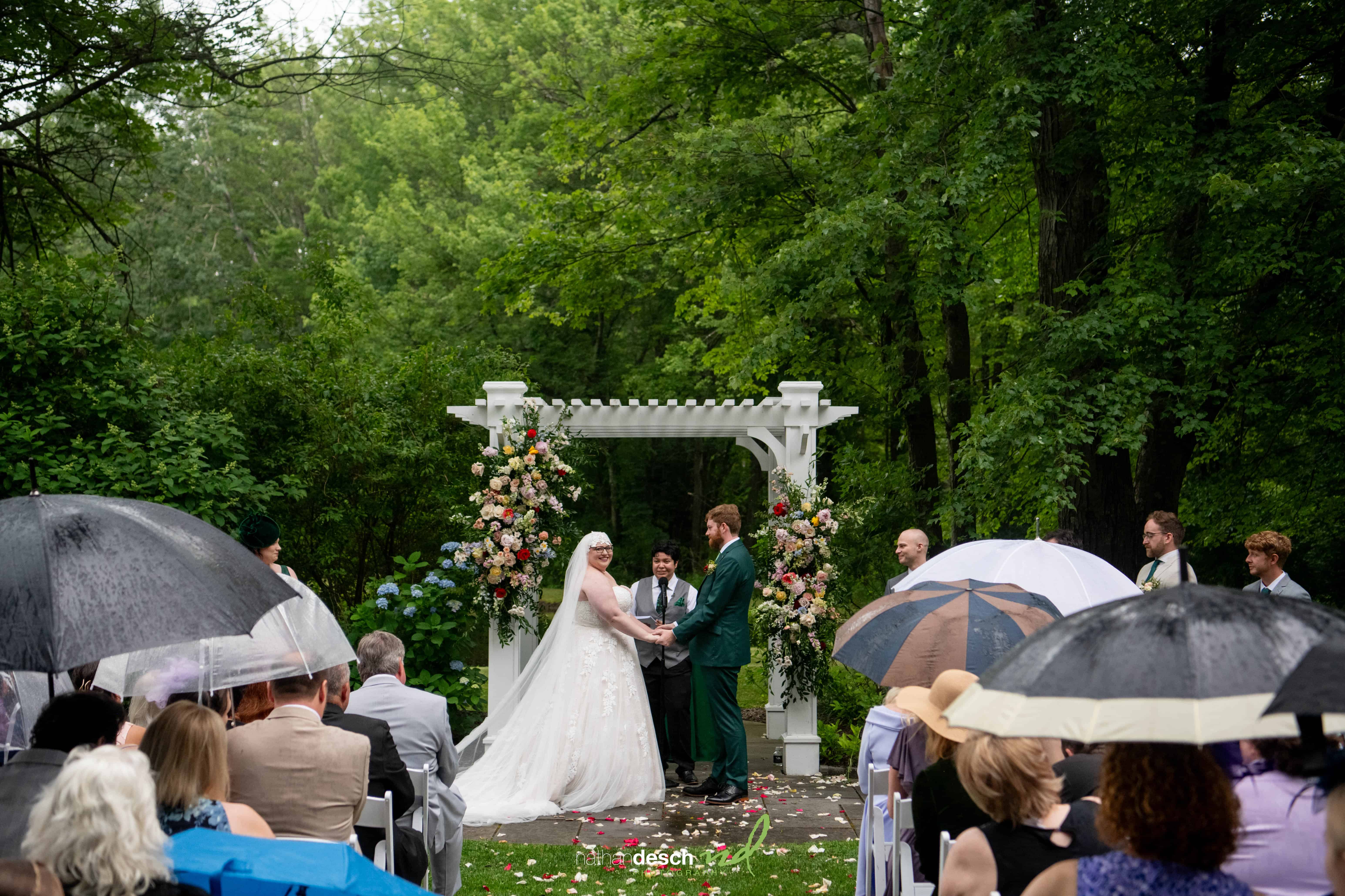 Bride and groom hold hands under floral arch at rainy outdoor wedding, guests with umbrellas, surrounded by lush green trees.