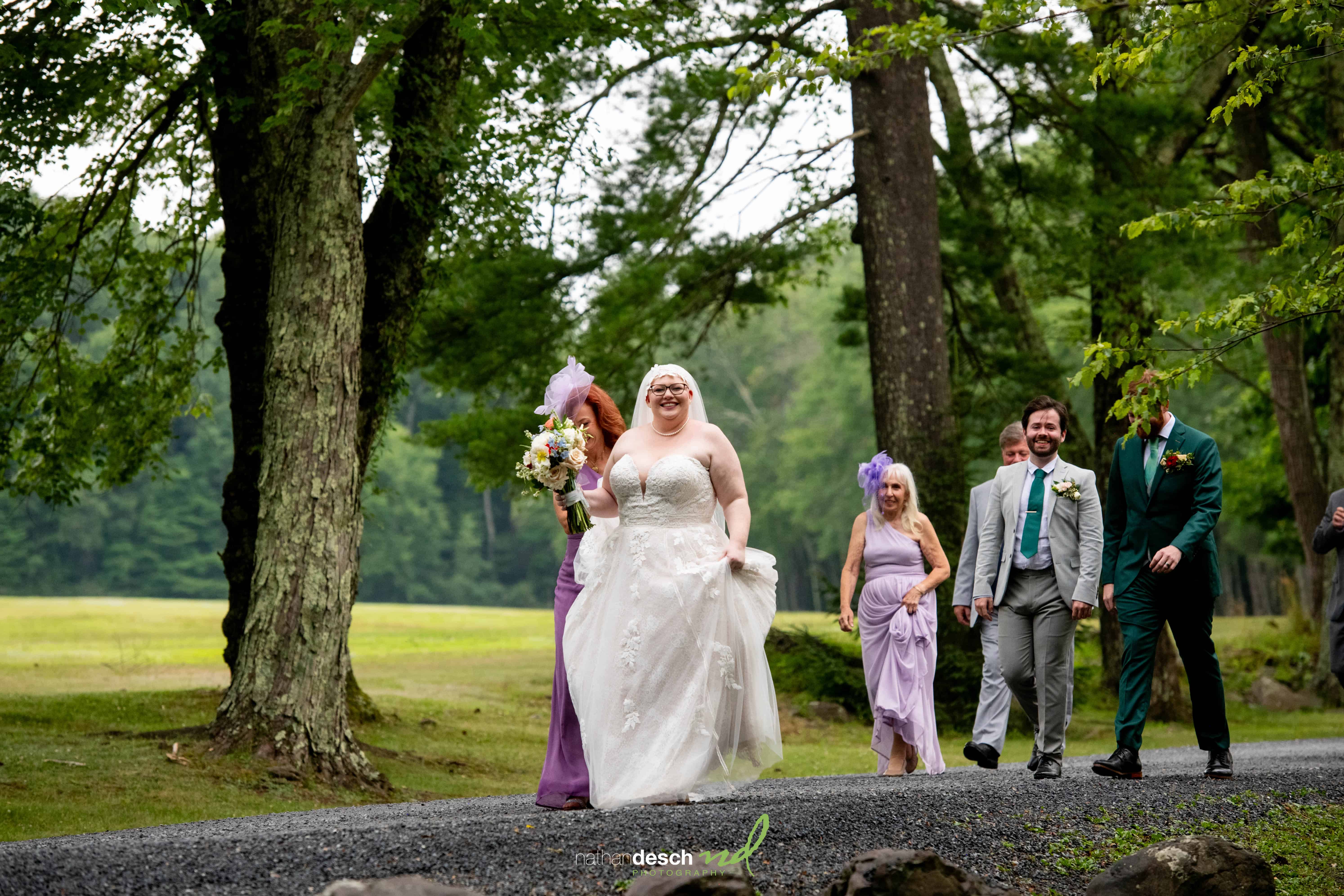 Bride in white dress walks with bridesmaids in lavender and groomsmen on gravel path during Jake and Erika’s outdoor wedding.