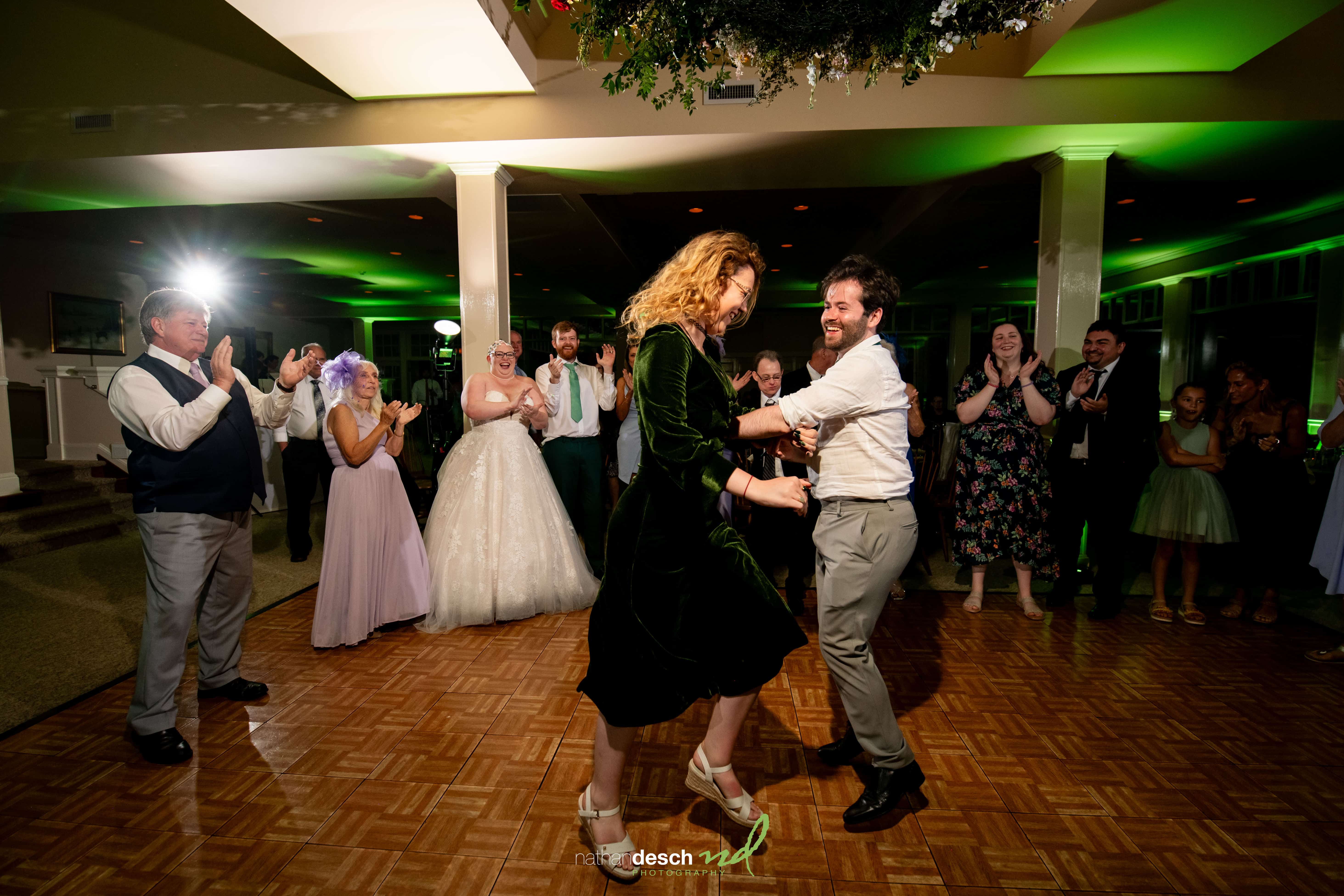 Joyful bride and groom dance on wooden floor, surrounded by smiling guests in formal wear at warmly lit wedding reception.