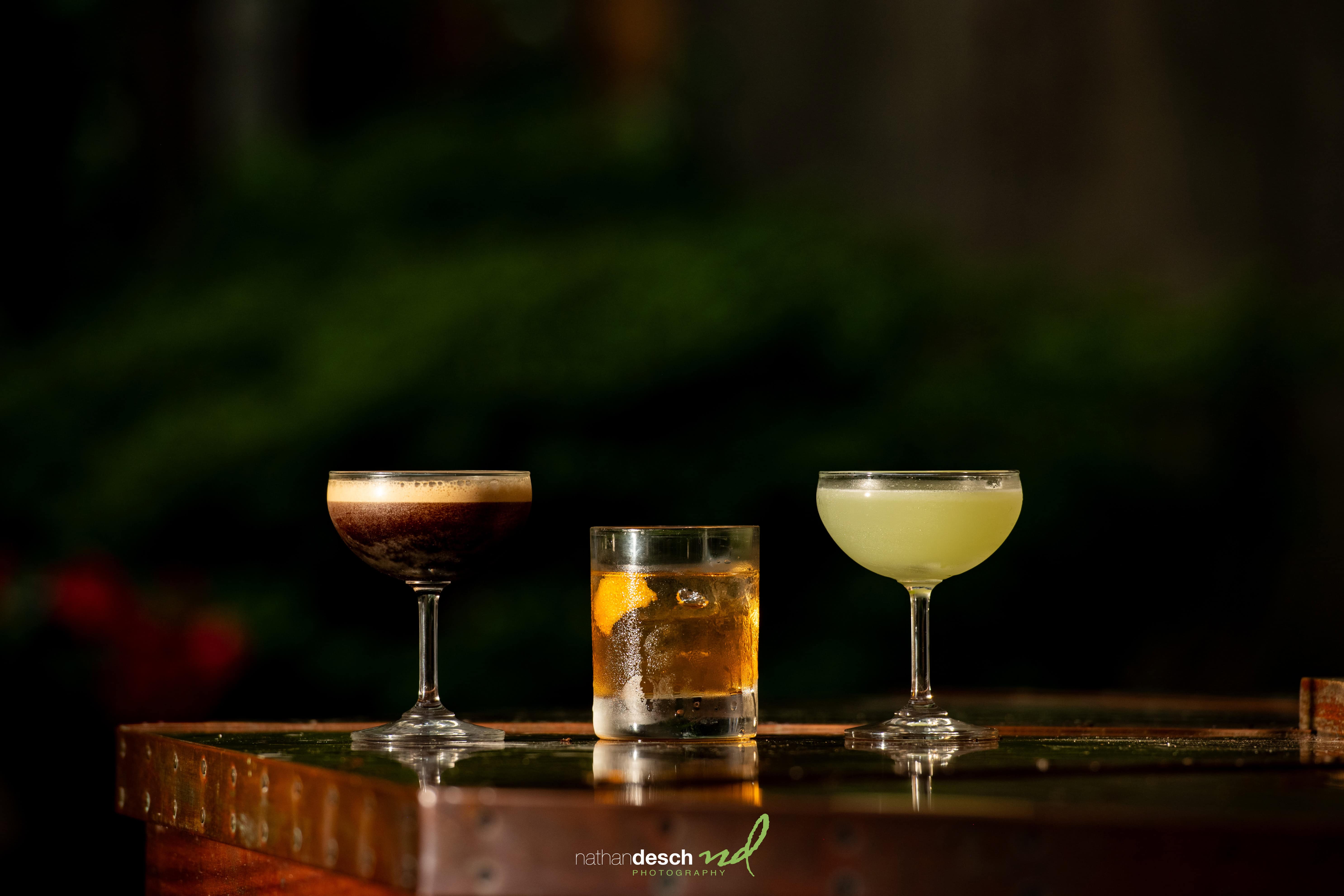 Three wedding cocktails—a dark espresso martini, whiskey on ice, and a green sour—on a rustic wooden bar with greenery.