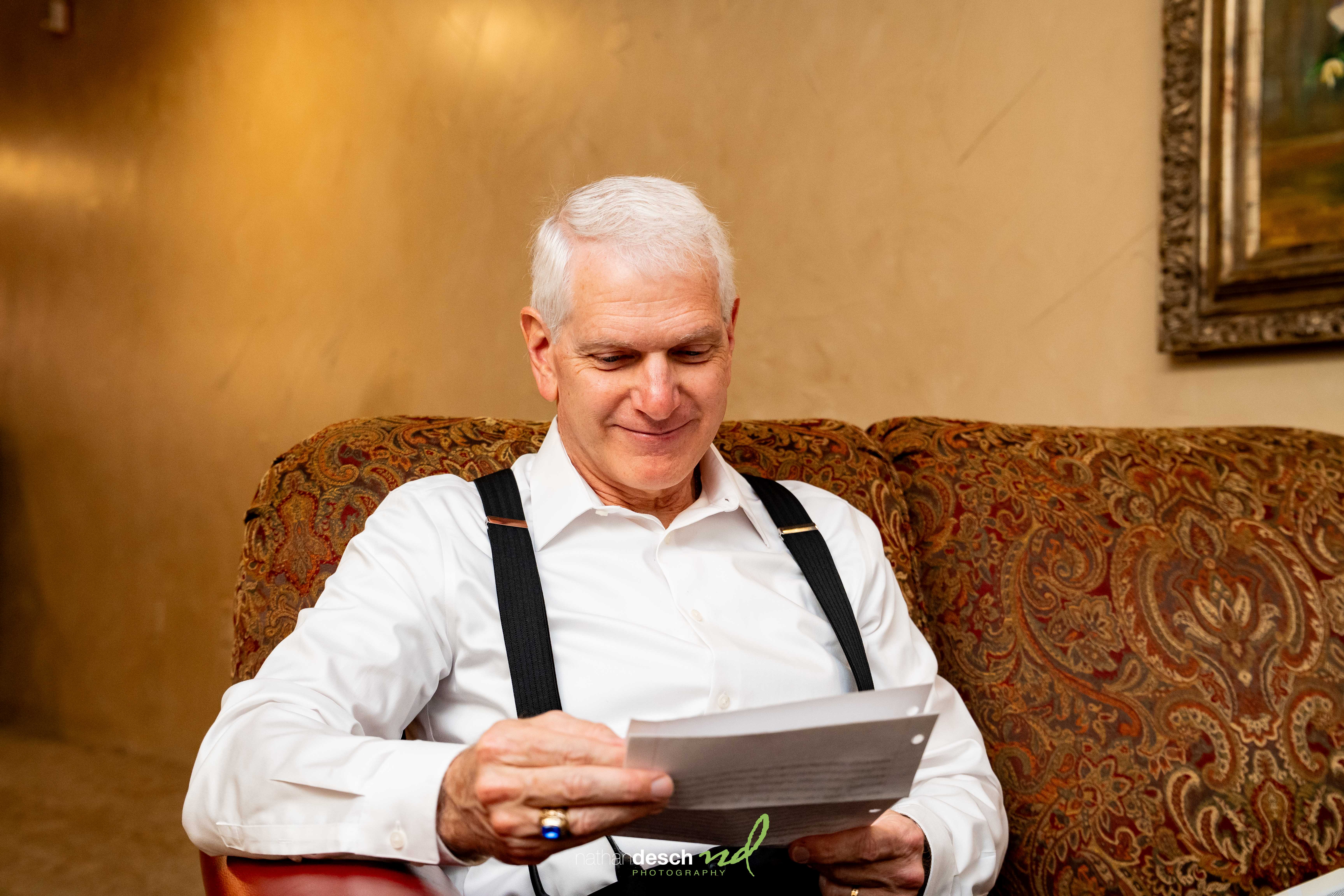 Older man with white hair in dress shirt and suspenders smiling while reading at Inn at Leola Village wedding reception.