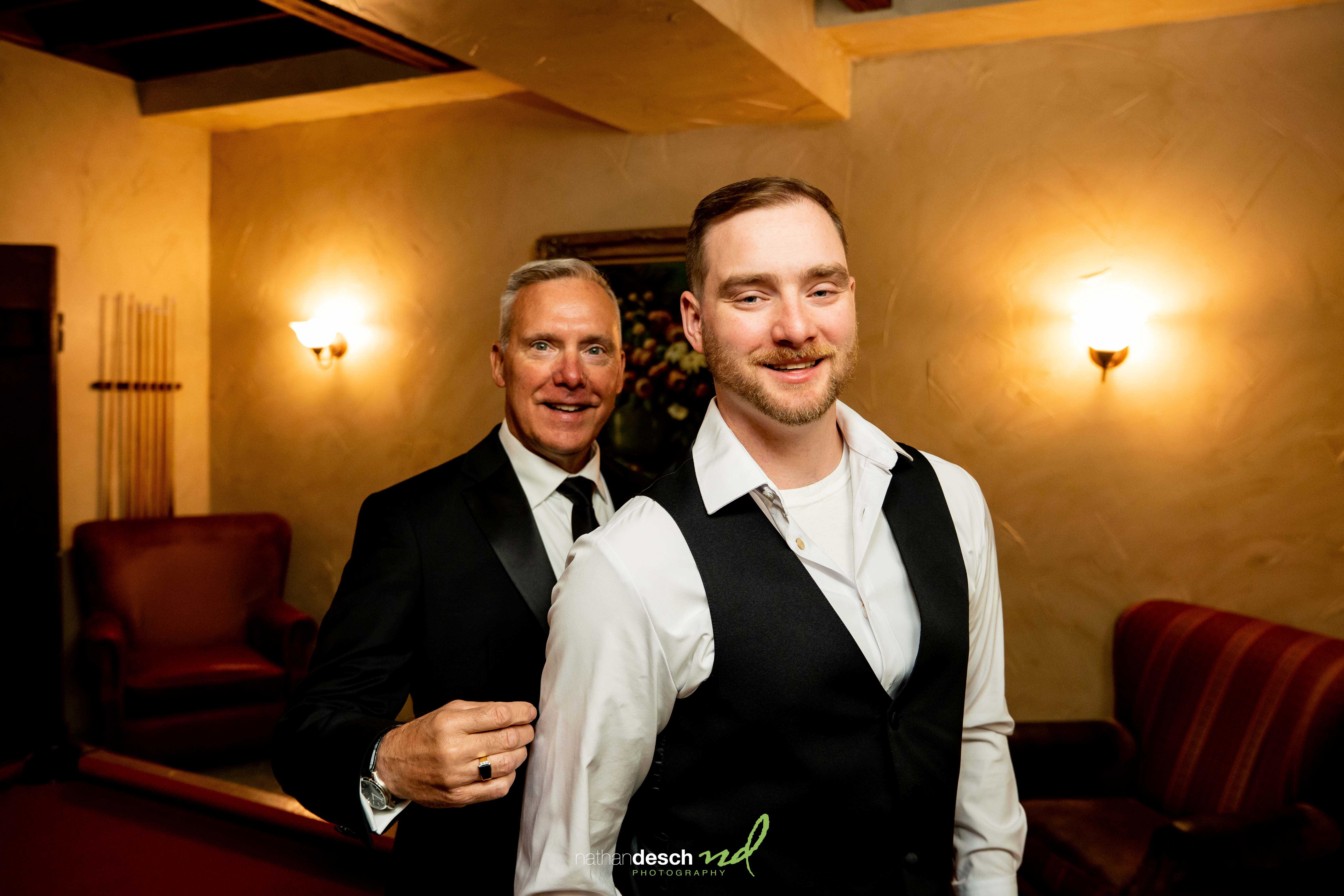 Groom smiling as partner adjusts his vest in warmly lit room at The Inn at Leola Village before their wedding ceremony.