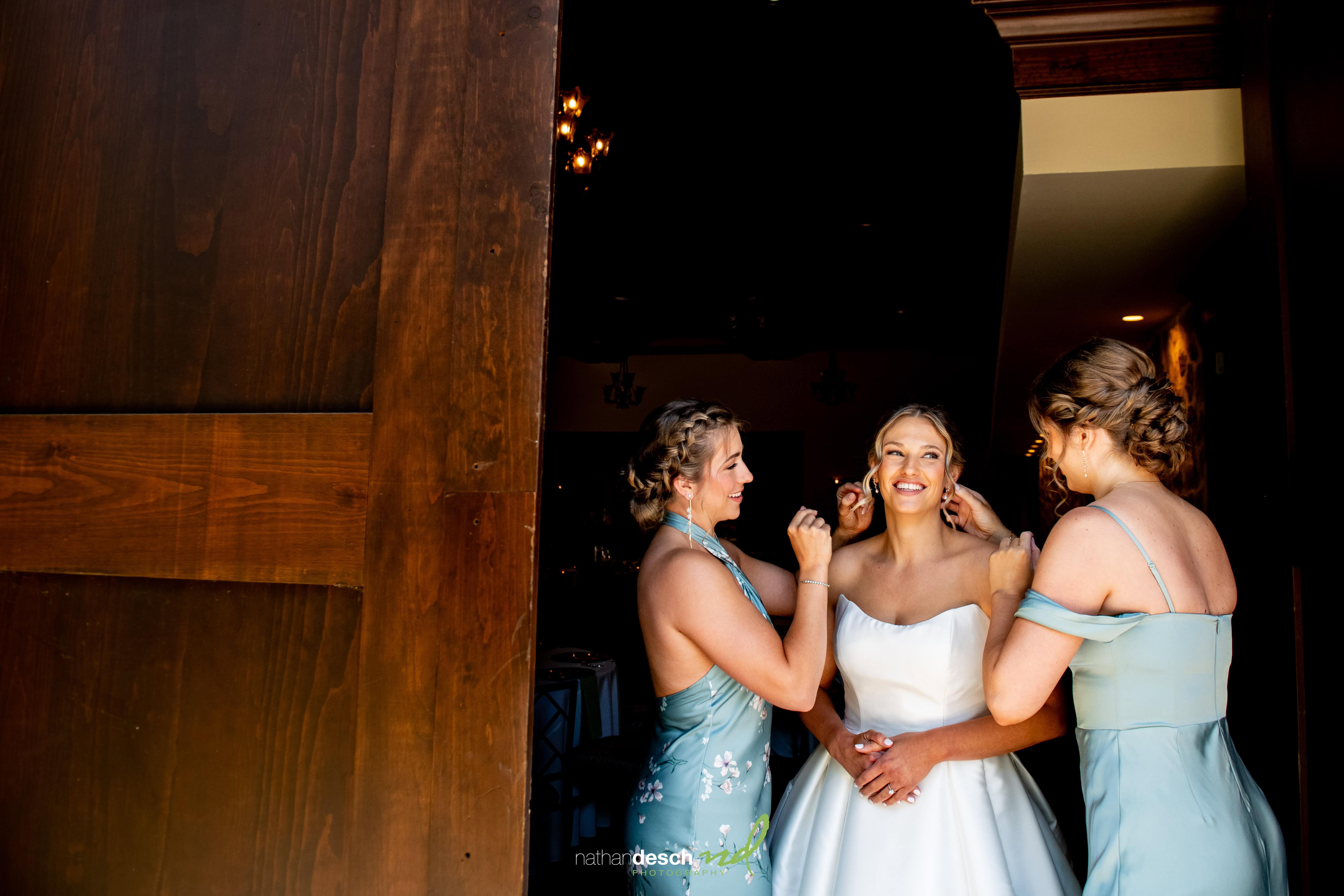 Bride smiling with two bridesmaids in light blue dresses getting ready by a wooden door at the Inn at Leola Village.