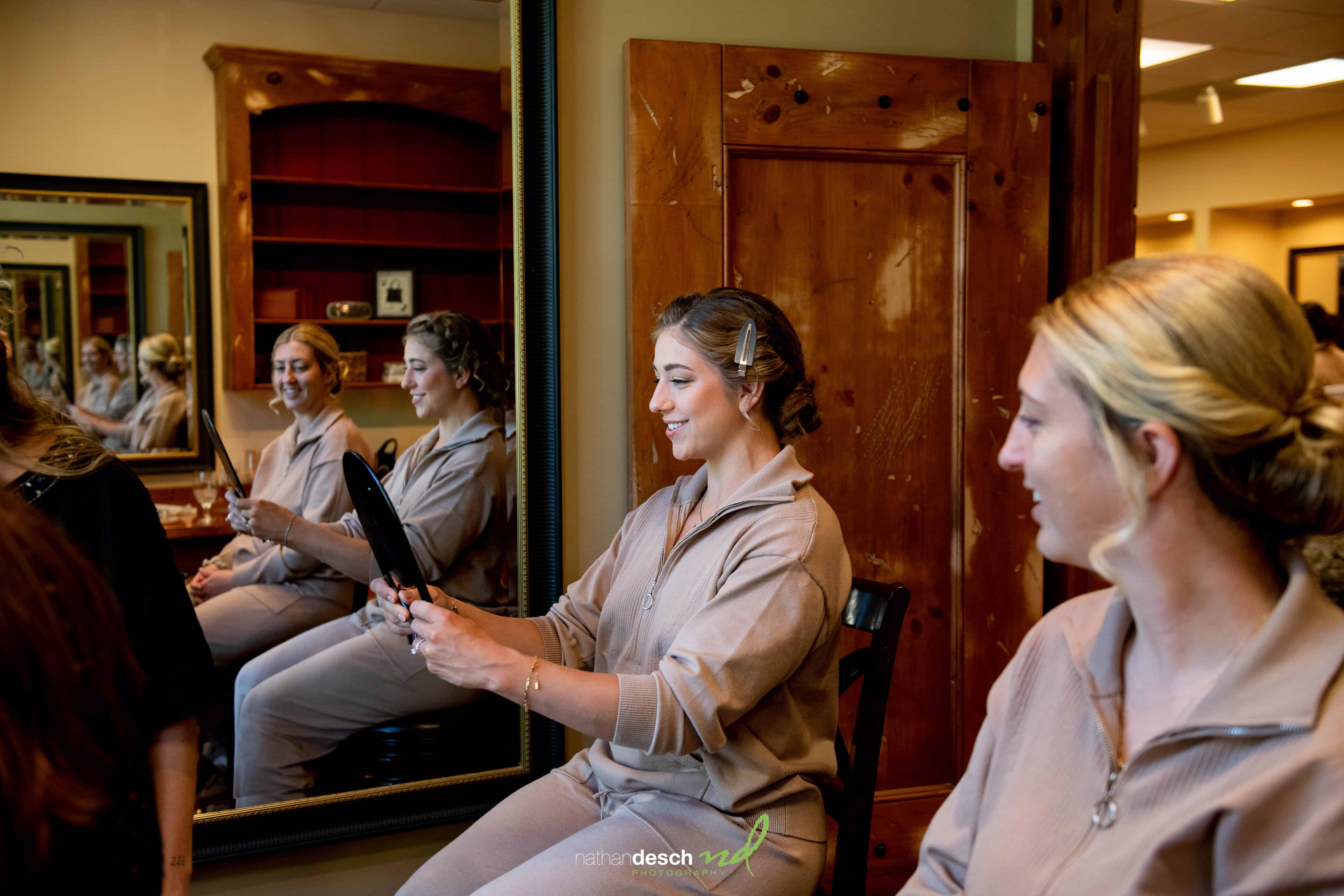 Bride in tan outfit smiles holding mirror at The Inn at Leola Village before elegant wedding; bridesmaid reflected nearby.