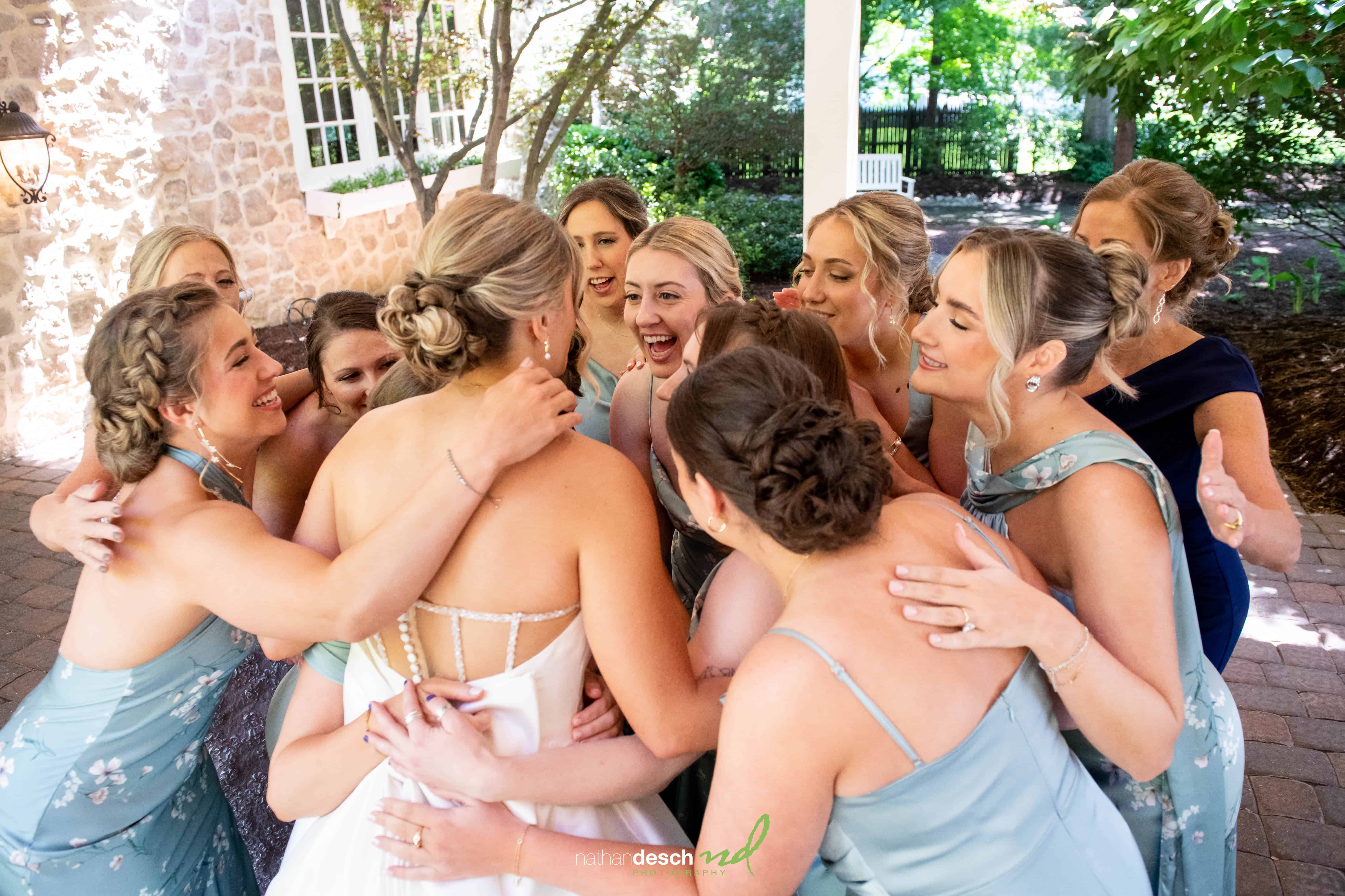 Bridesmaids in blue hug bride in white gown outdoors at Inn at Leola Village, surrounded by greenery and stone walls.