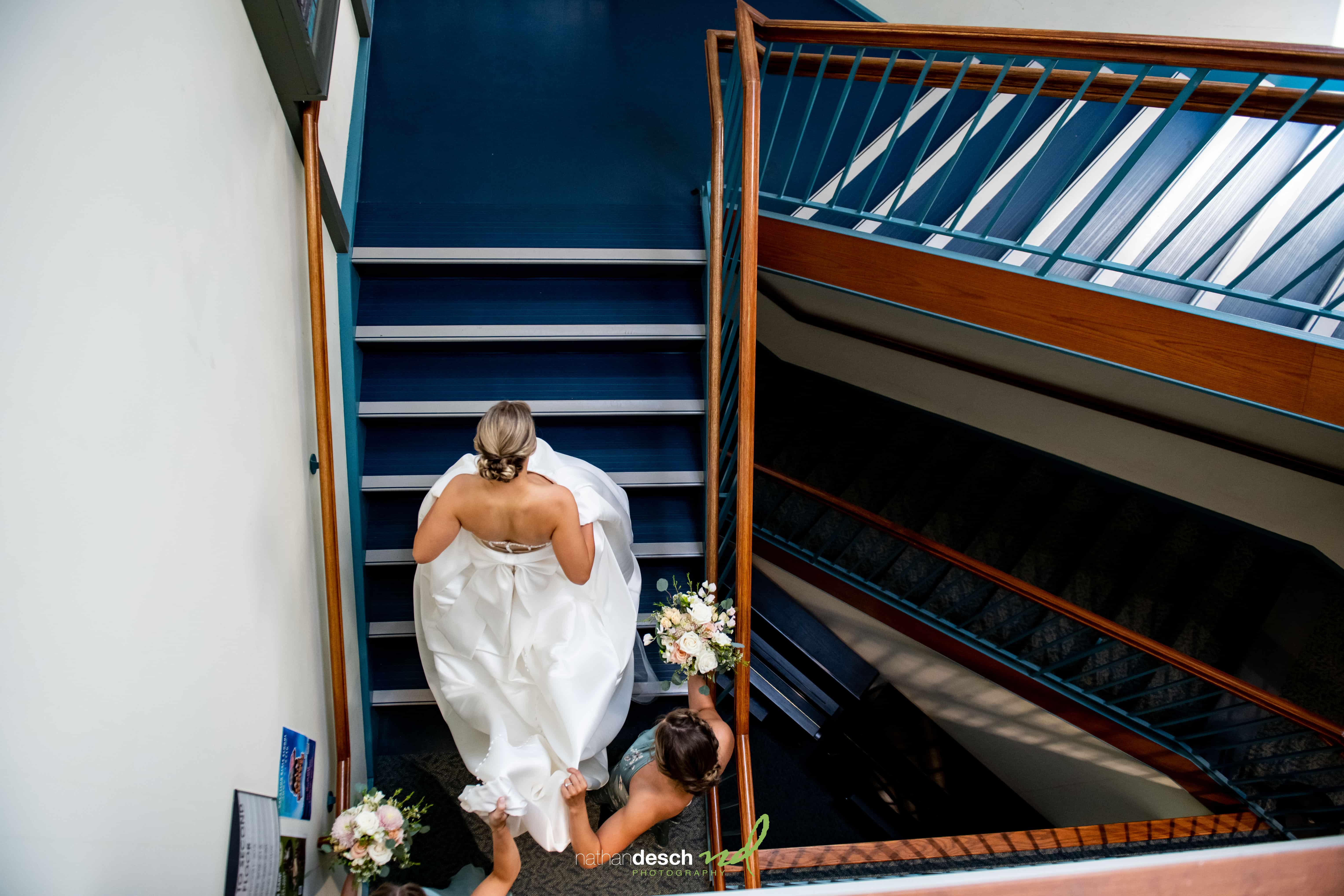 Bride in white dress walks up blue stairs at The Inn at Leola Village, followed by bridesmaid holding flowers on wedding day.