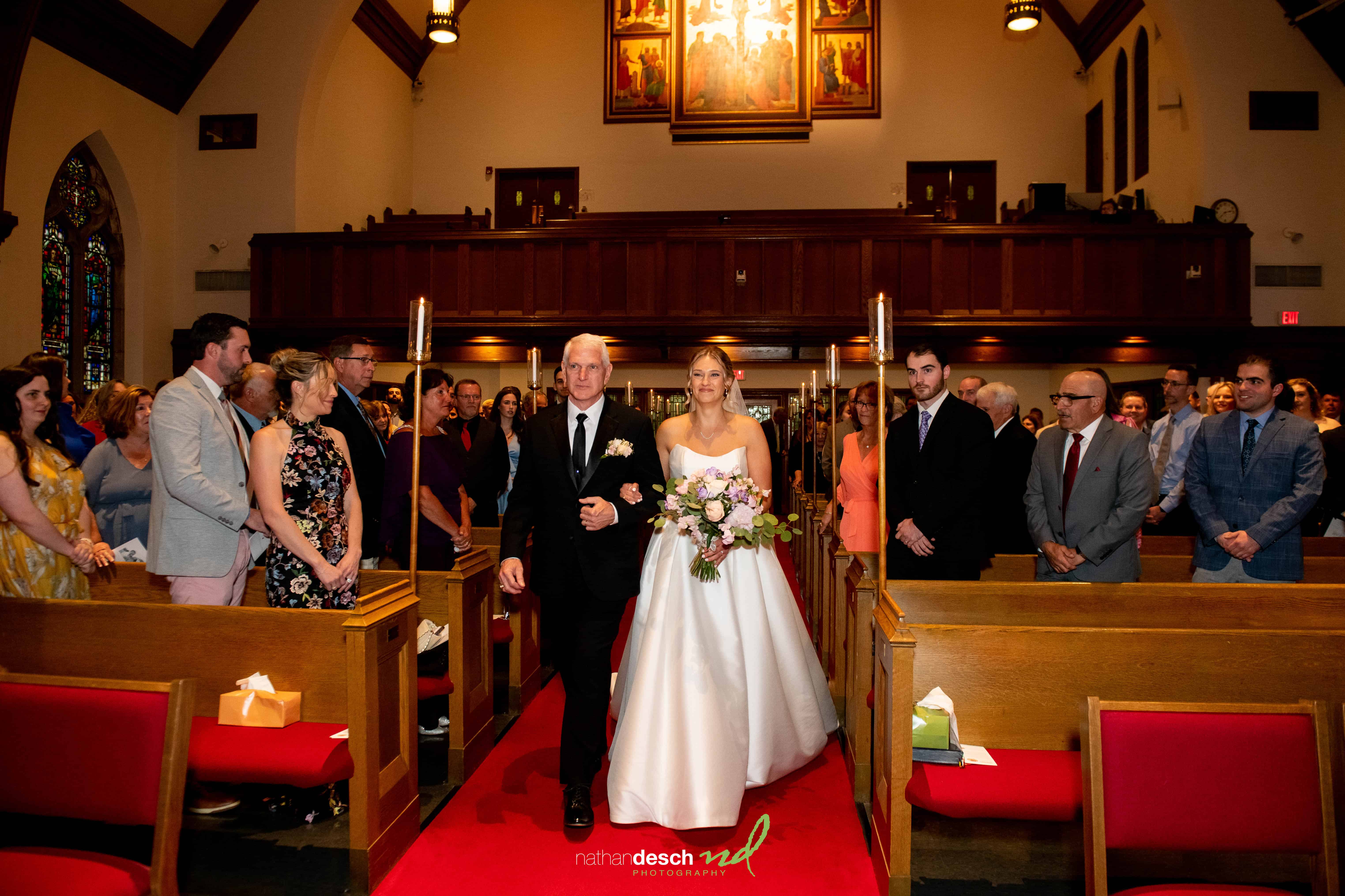 Bride in white gown walks down church aisle with groom, holding bouquet, surrounded by guests and stained glass windows.