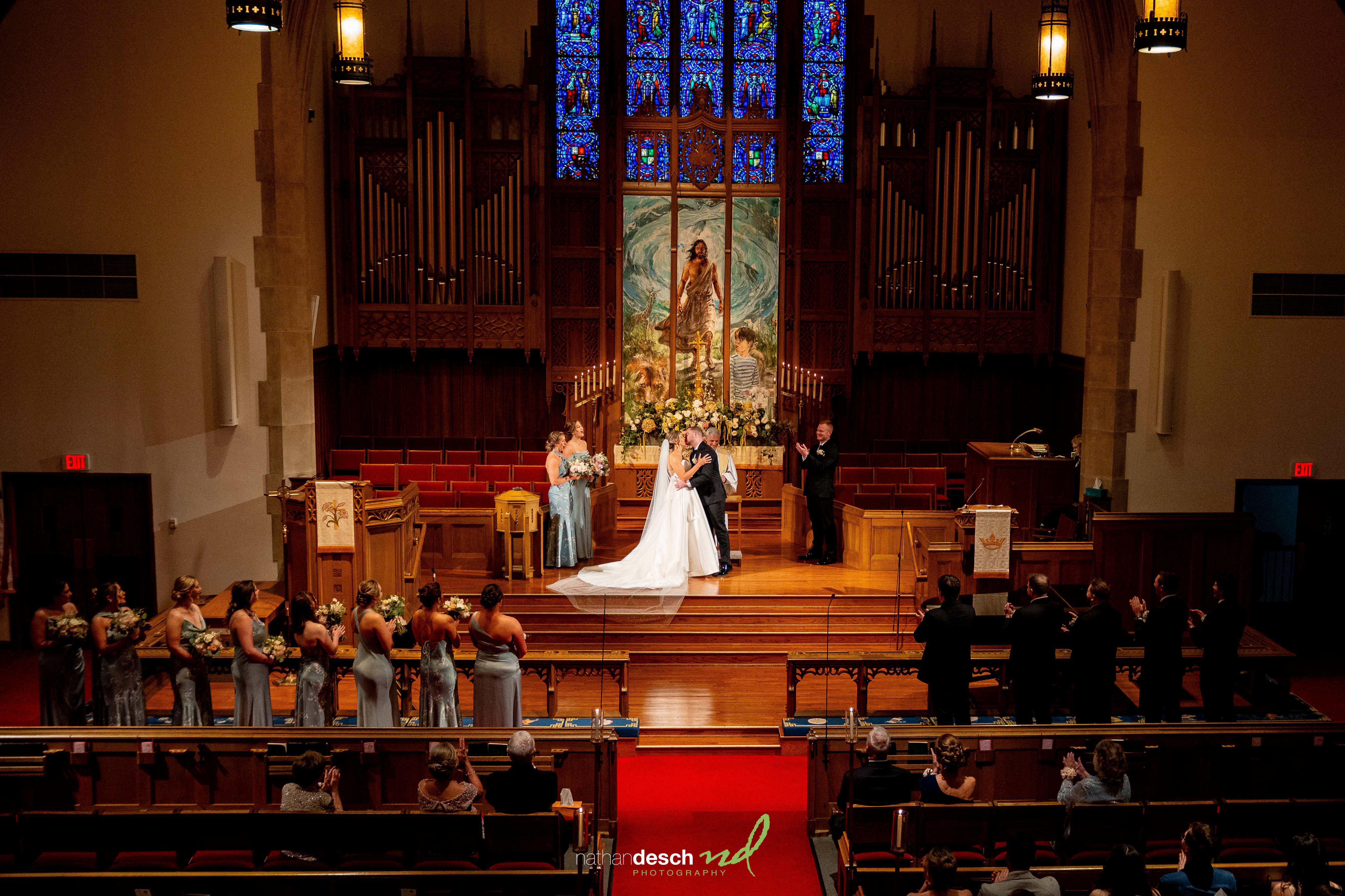 Bride and groom share first kiss at church altar with stained glass, surrounded by loved ones at elegant Inn at Leola Village wedding.