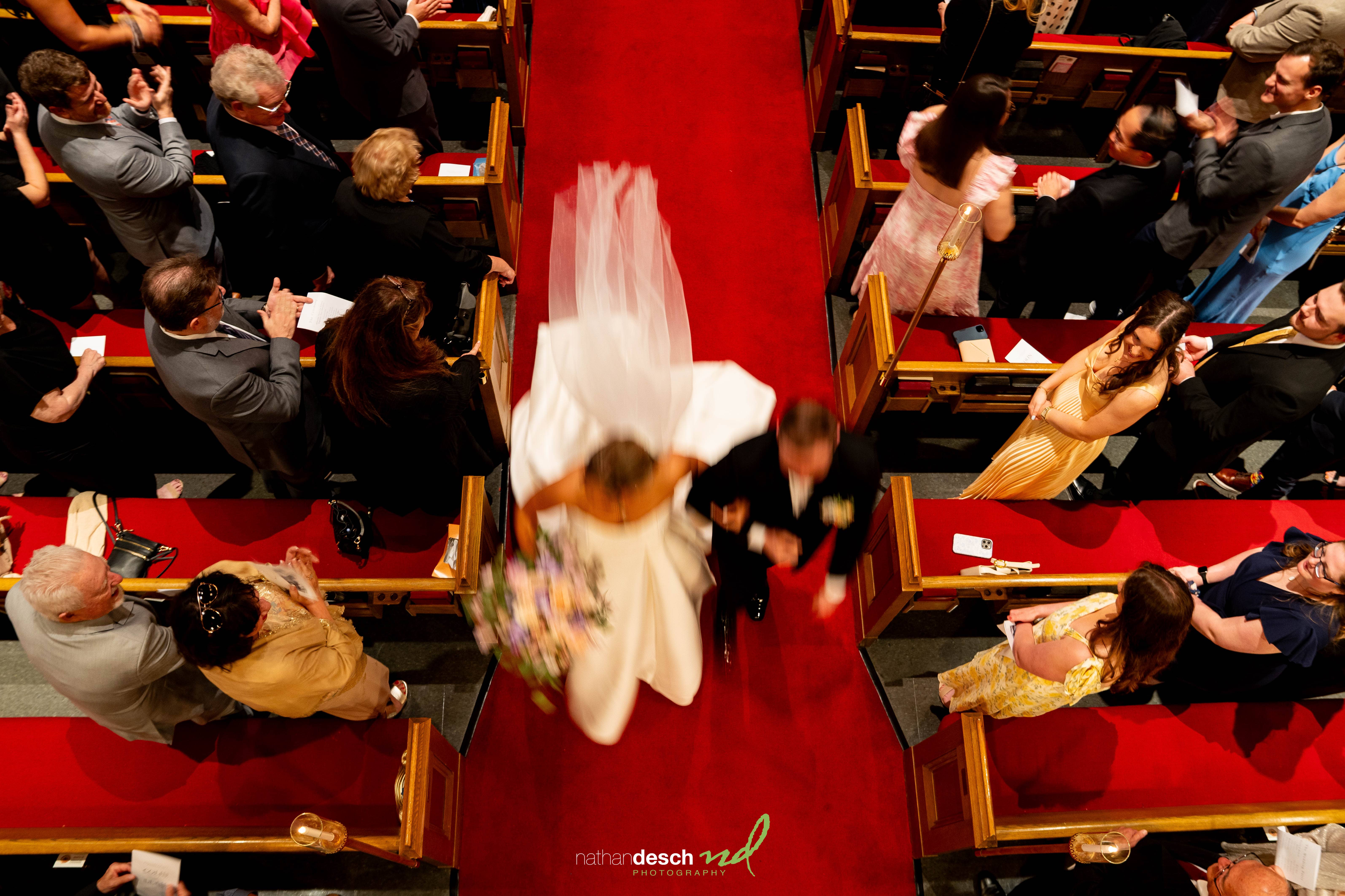 Bride and groom walk down the aisle at The Inn at Leola Village as guests stand and applaud, with bride’s veil trailing behind.