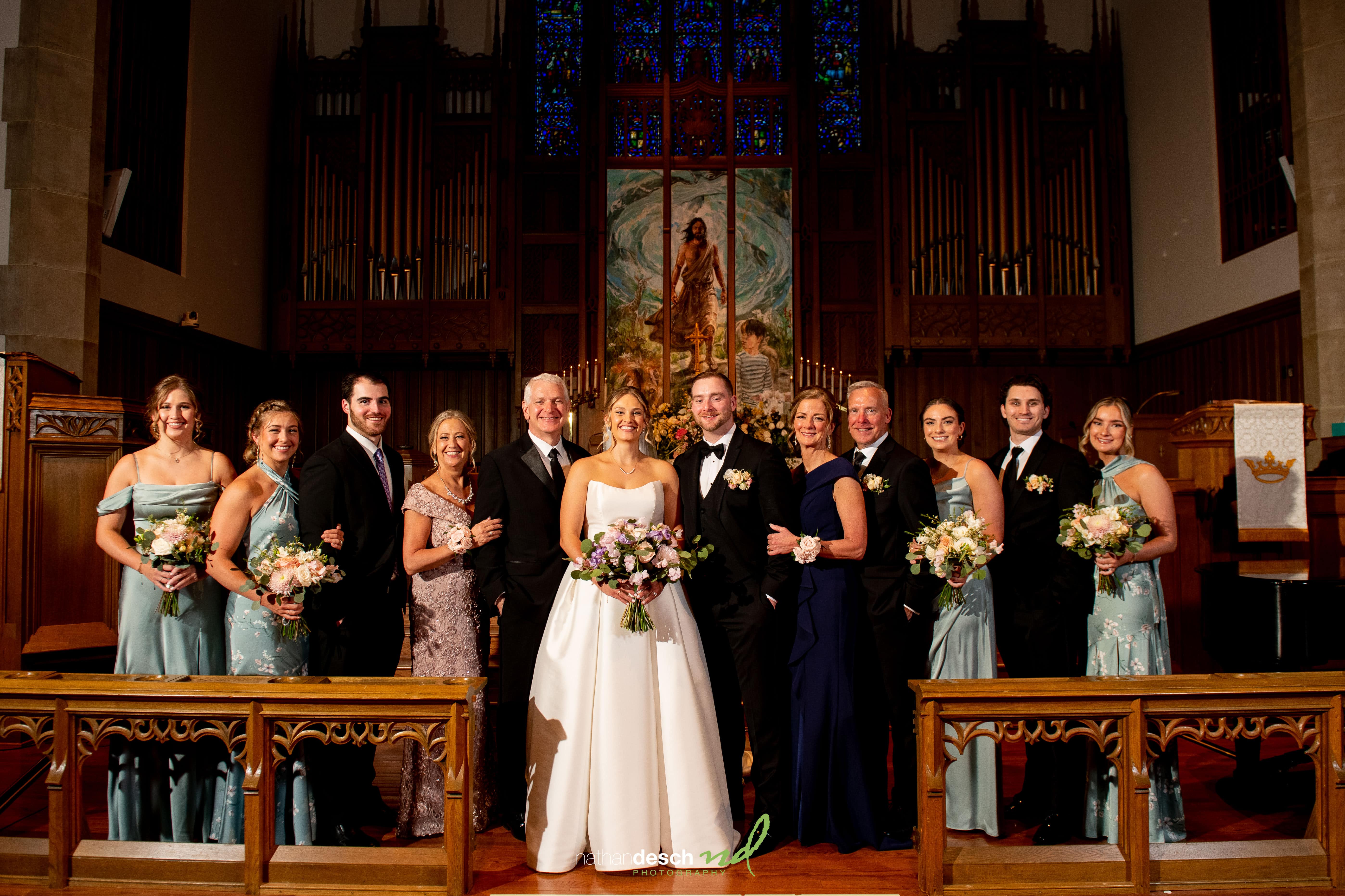 Wedding party posing in a church at The Inn at Leola Village, with bride and groom center, bridesmaids in blue, groomsmen in black.