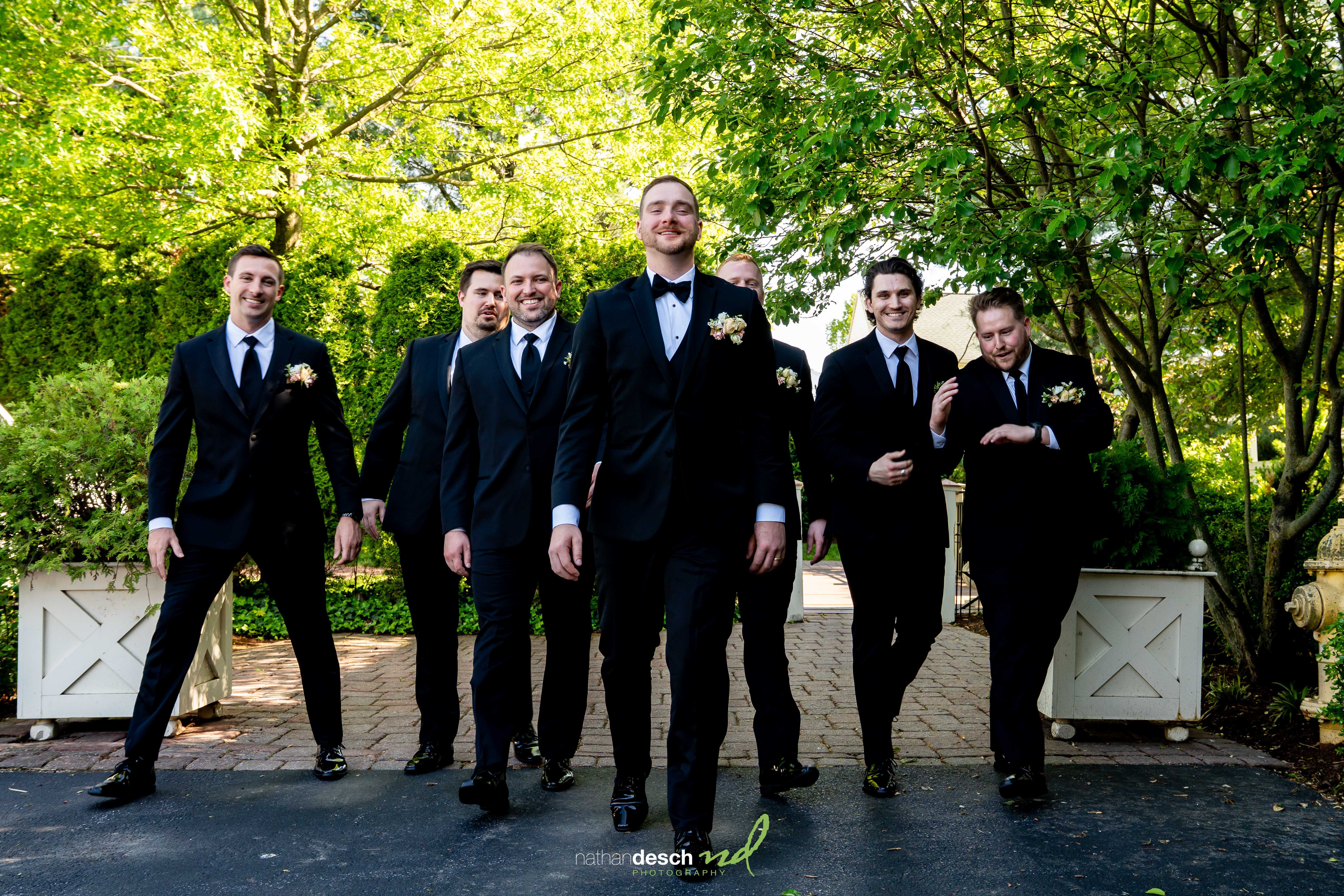 Groomsmen in black suits smiling and walking outdoors at The Inn at Leola Village, surrounded by lush greenery and trees.