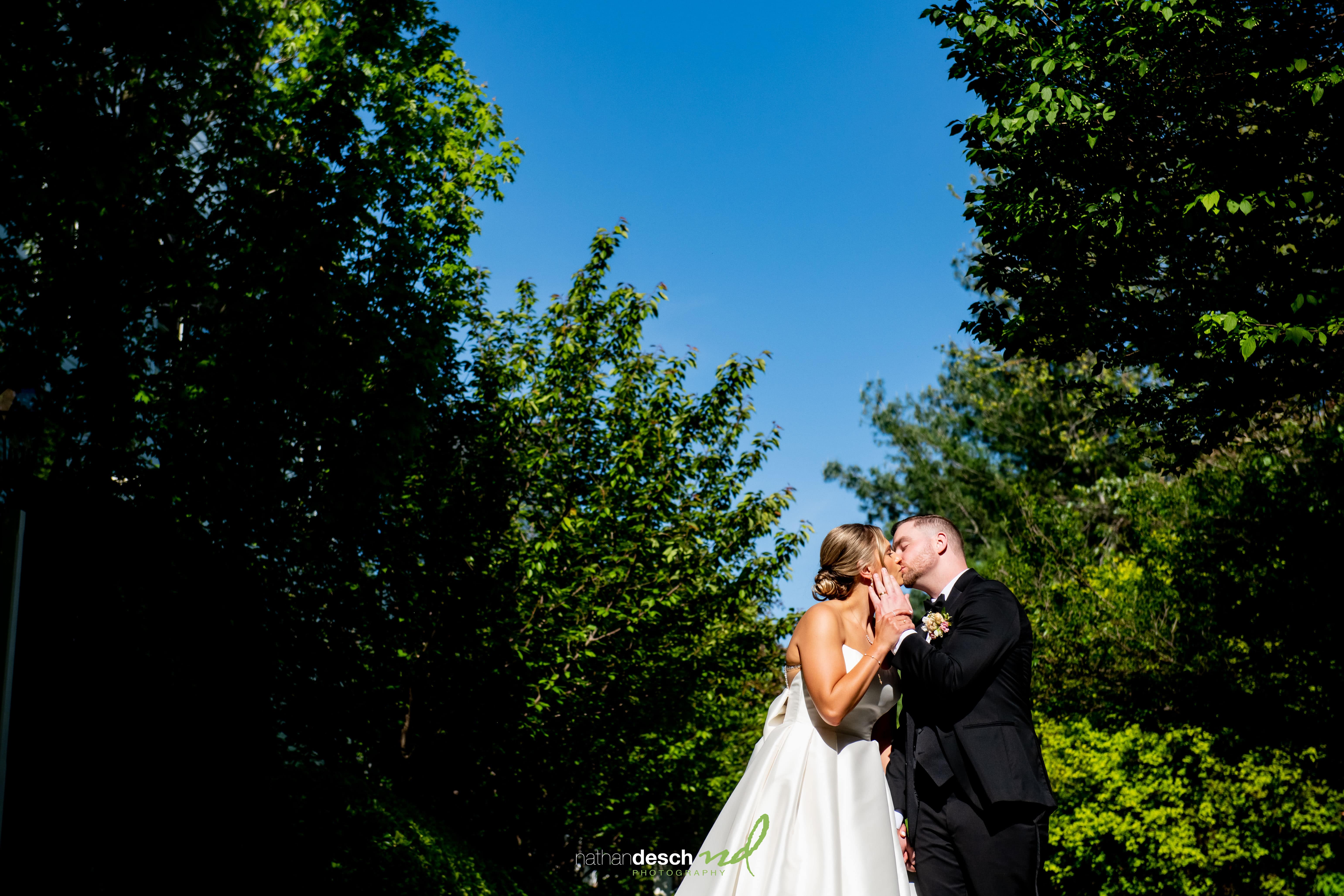 Bride and groom kiss outdoors at Inn at Leola Village, surrounded by lush greenery and blue sky on wedding day.