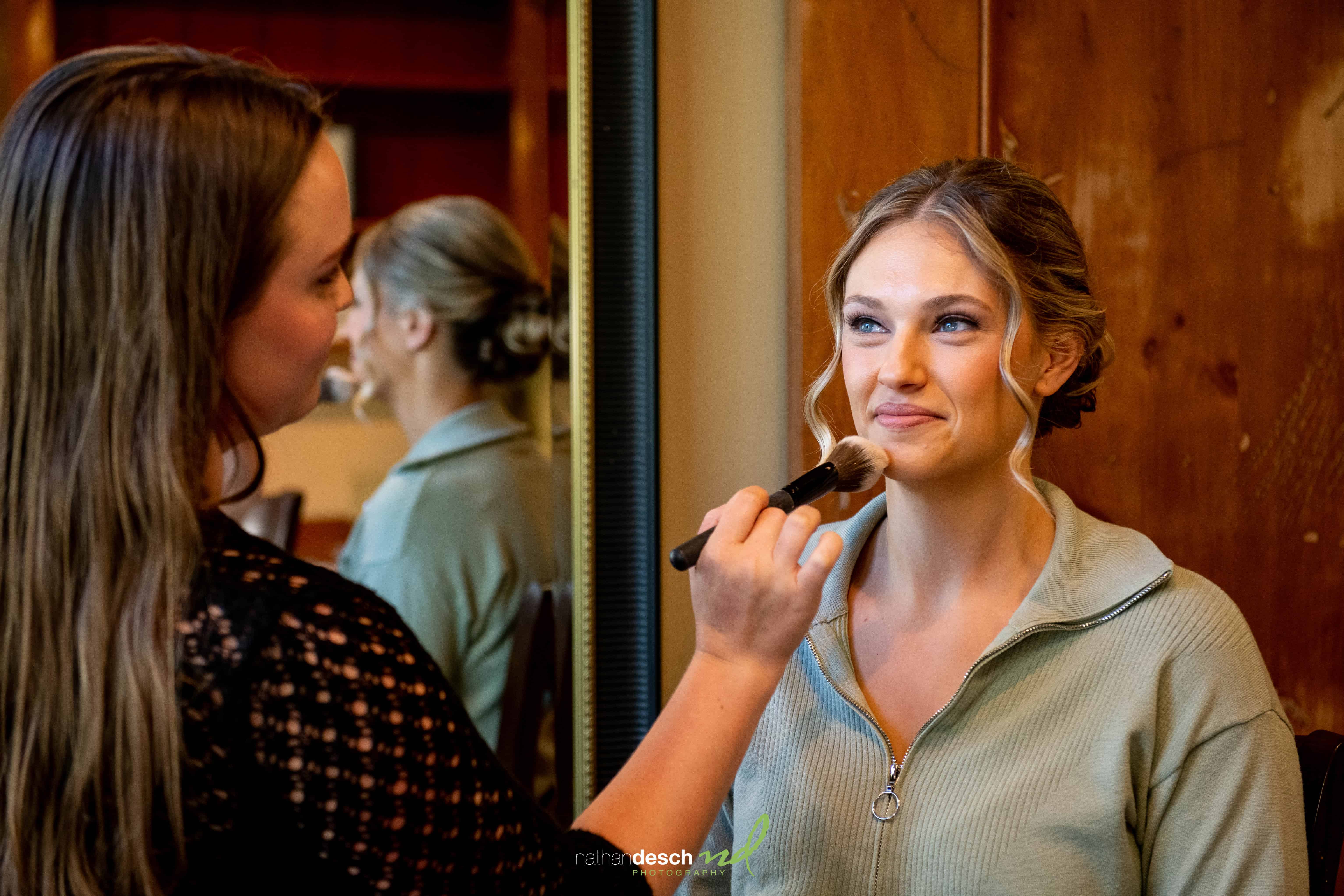Makeup artist applies blush to smiling bride in green zip-up top, preparing for wedding at Inn at Leola Village, warmly lit room.