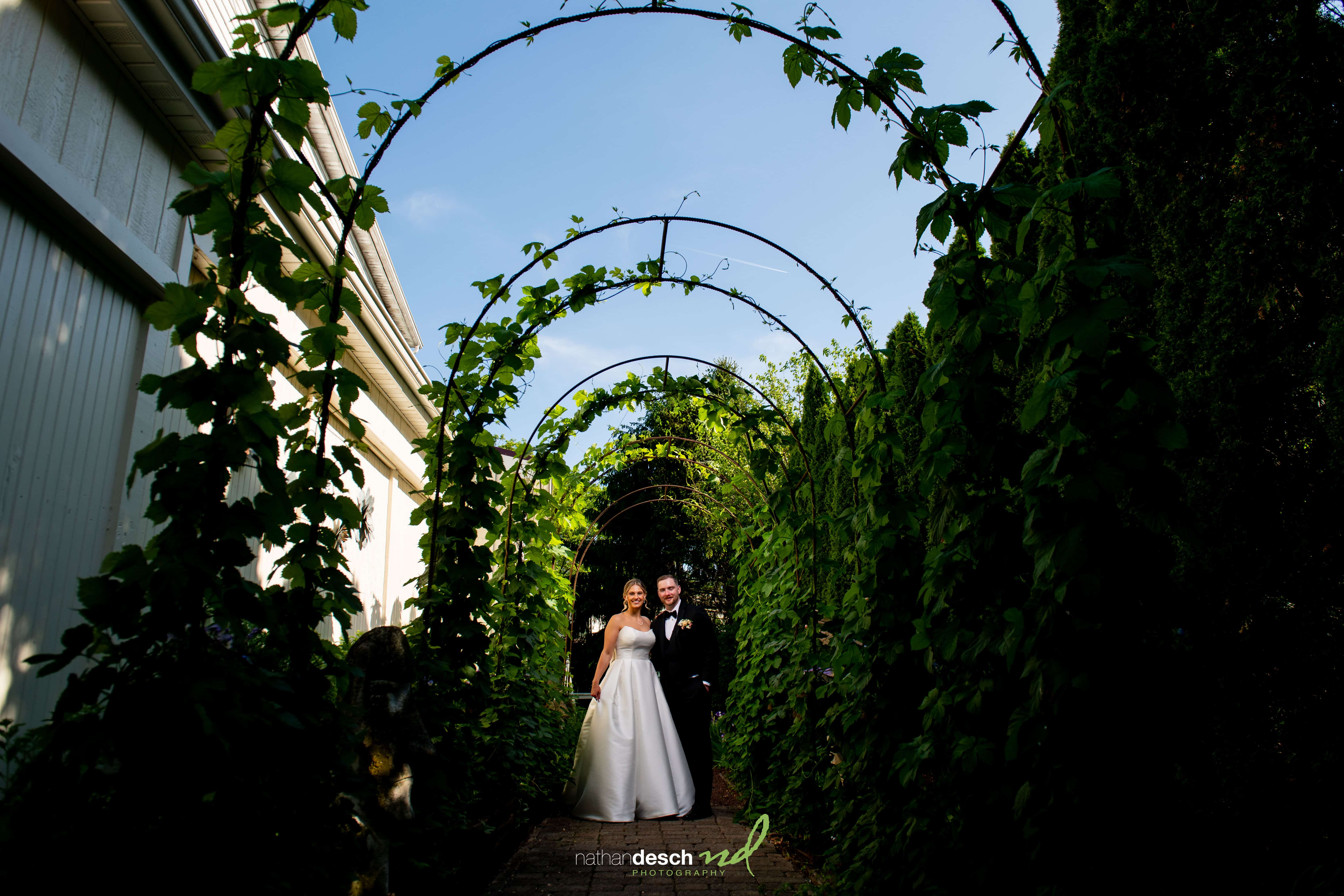 Bride and groom smiling under leafy archway at The Inn at Leola Village garden wedding, sunlight highlighting dress and tuxedo.