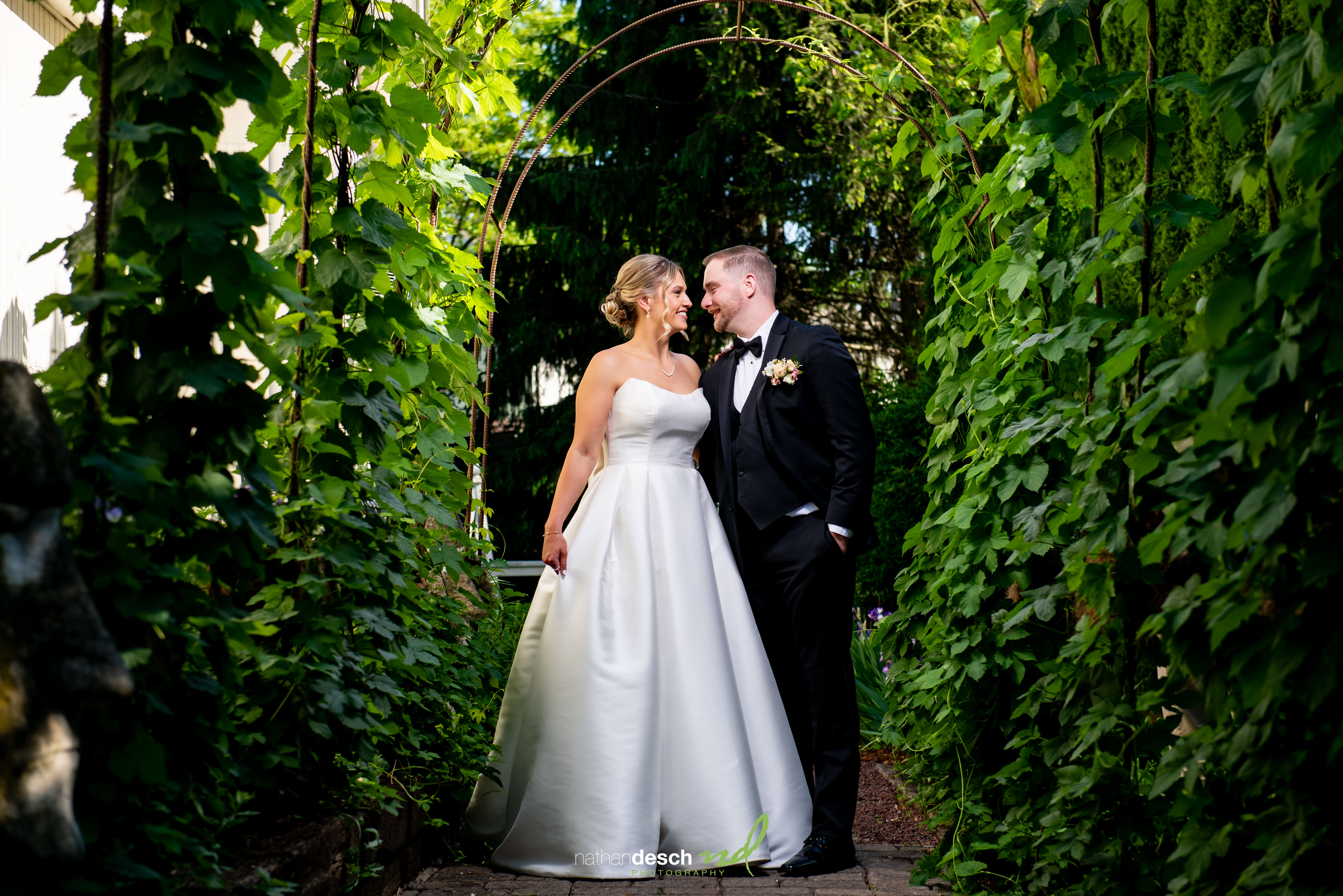 Bride and groom smiling under leafy garden archway at Inn at Leola Village wedding with sunlight shining through.