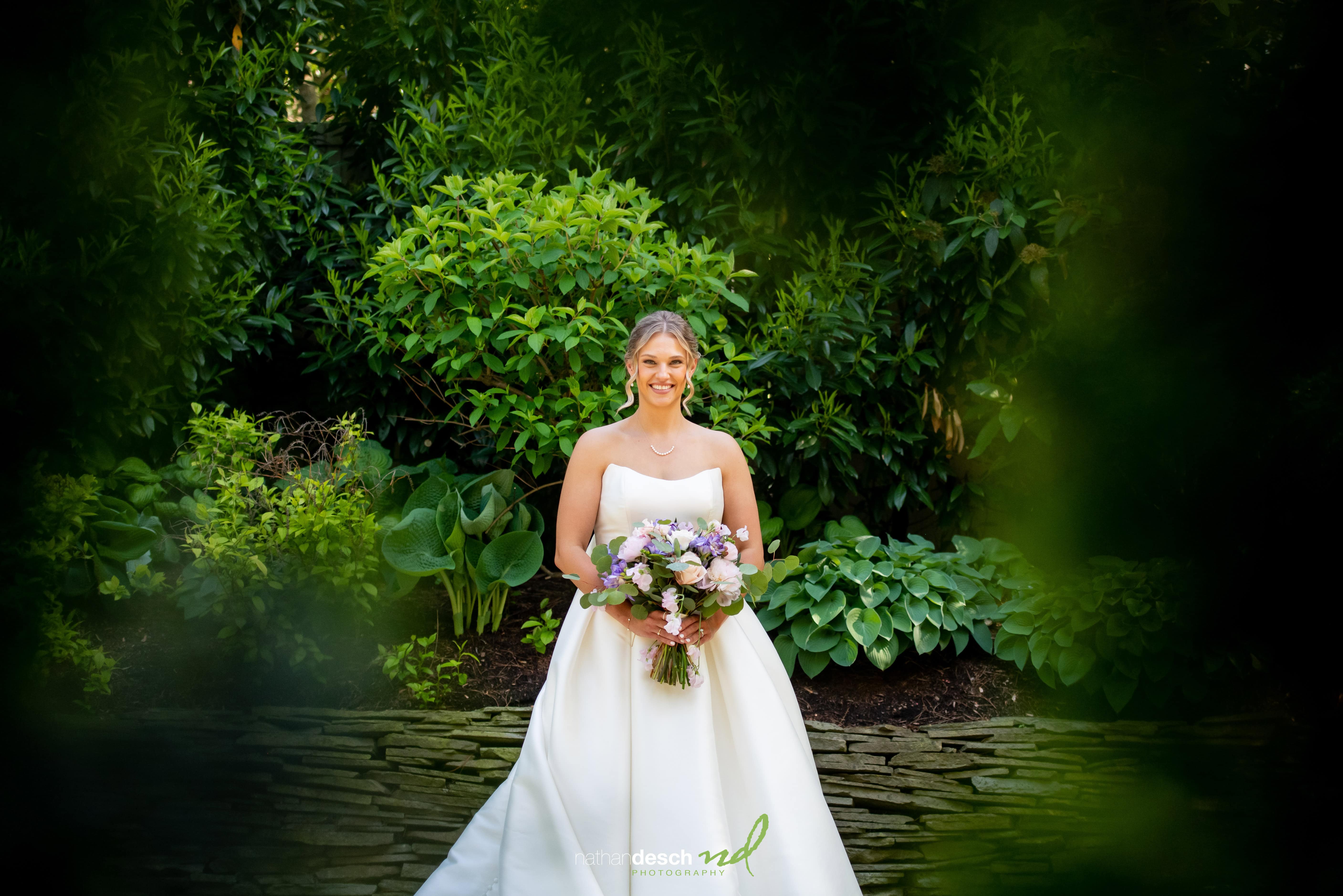 Bride in white dress smiling with bouquet at The Inn at Leola Village, surrounded by lush greenery and stone pavement in spring.