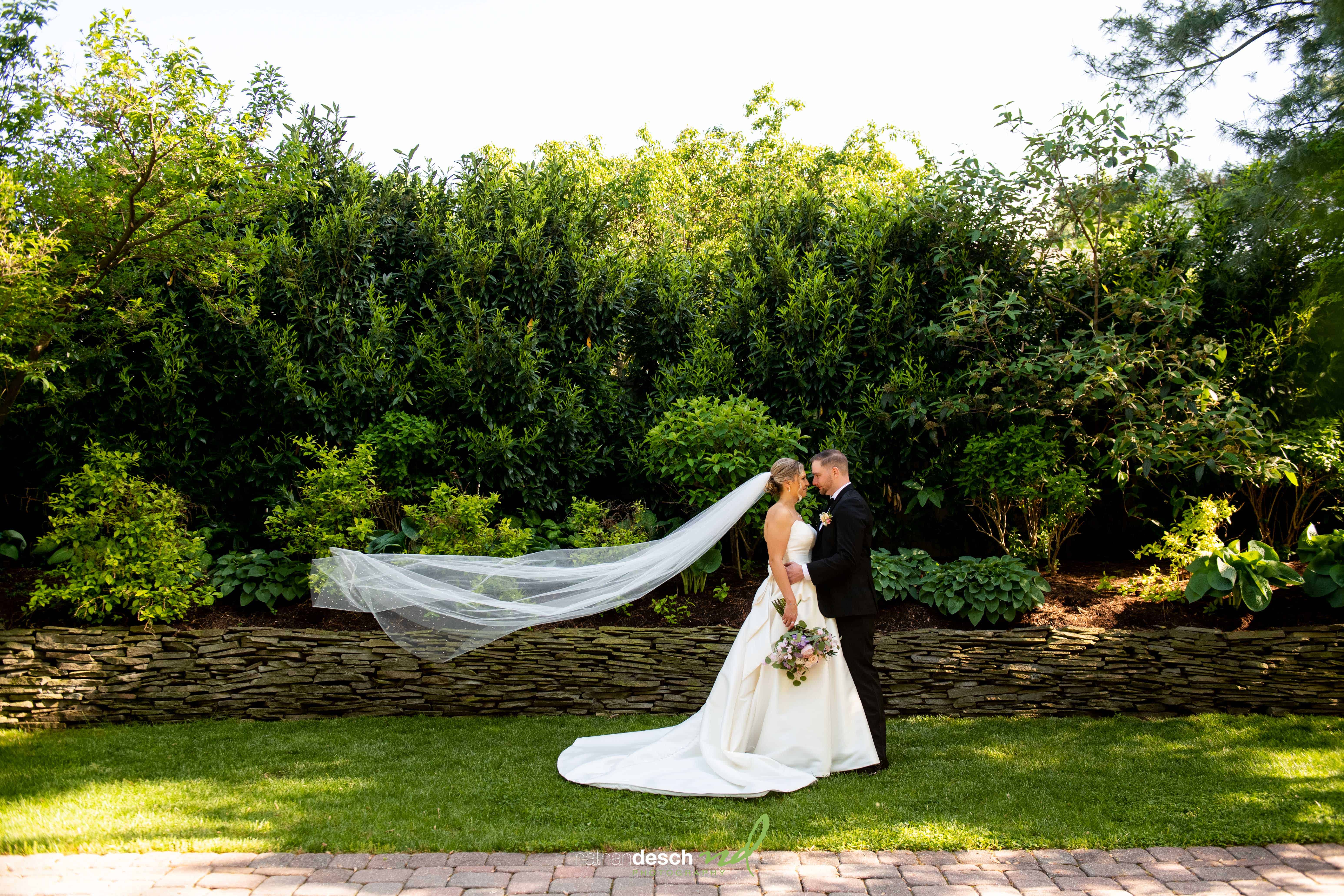Bride and groom kiss on green lawn at The Inn at Leola Village, sunlight on her flowing veil, bouquet in hand, lush hedge backdrop.