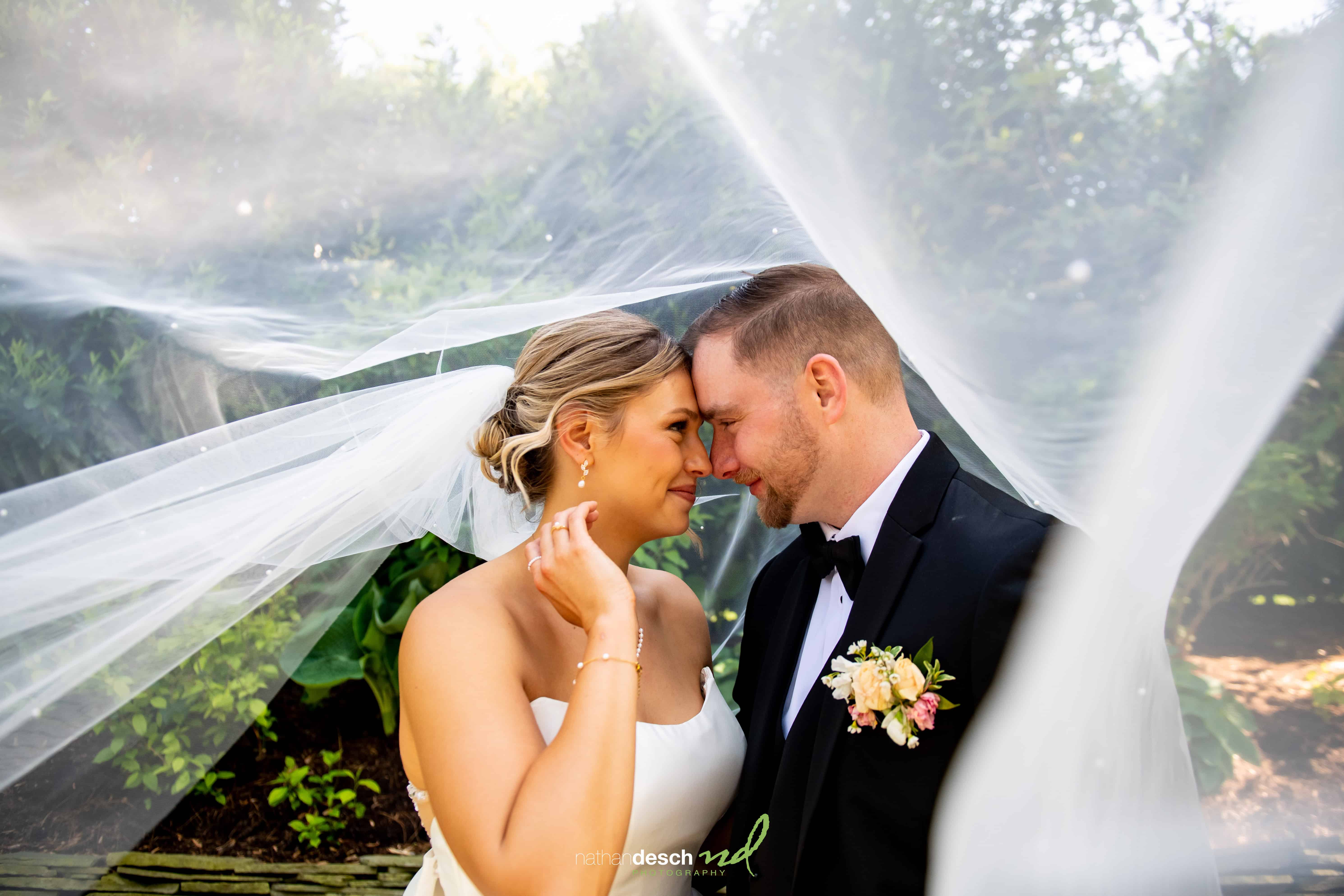 Bride and groom smiling under a sheer veil at The Inn at Leola Village, surrounded by lush greenery on their wedding day.