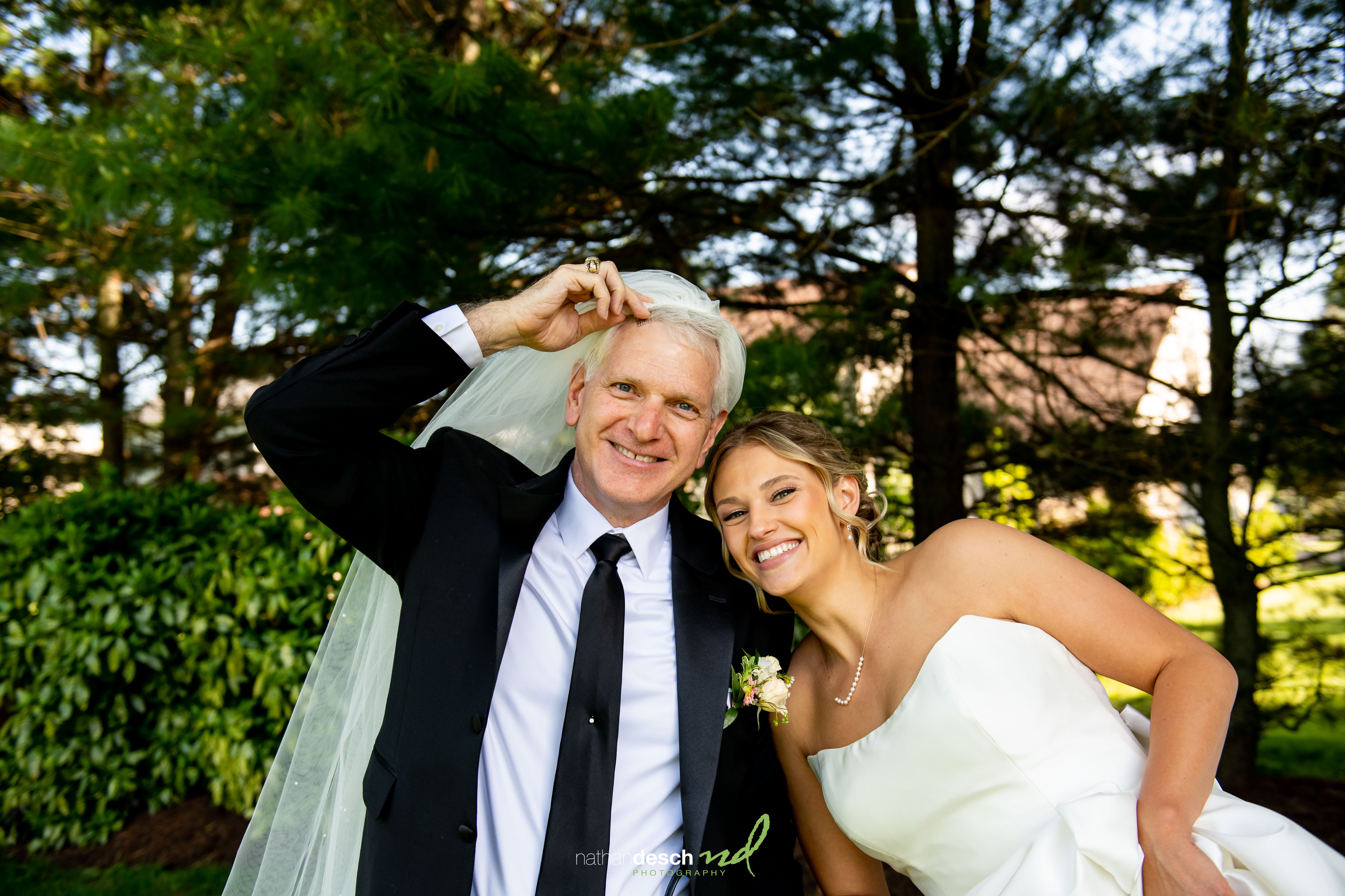 Groom in suit playfully wears bridal veil beside laughing bride outdoors, with lush green trees behind.