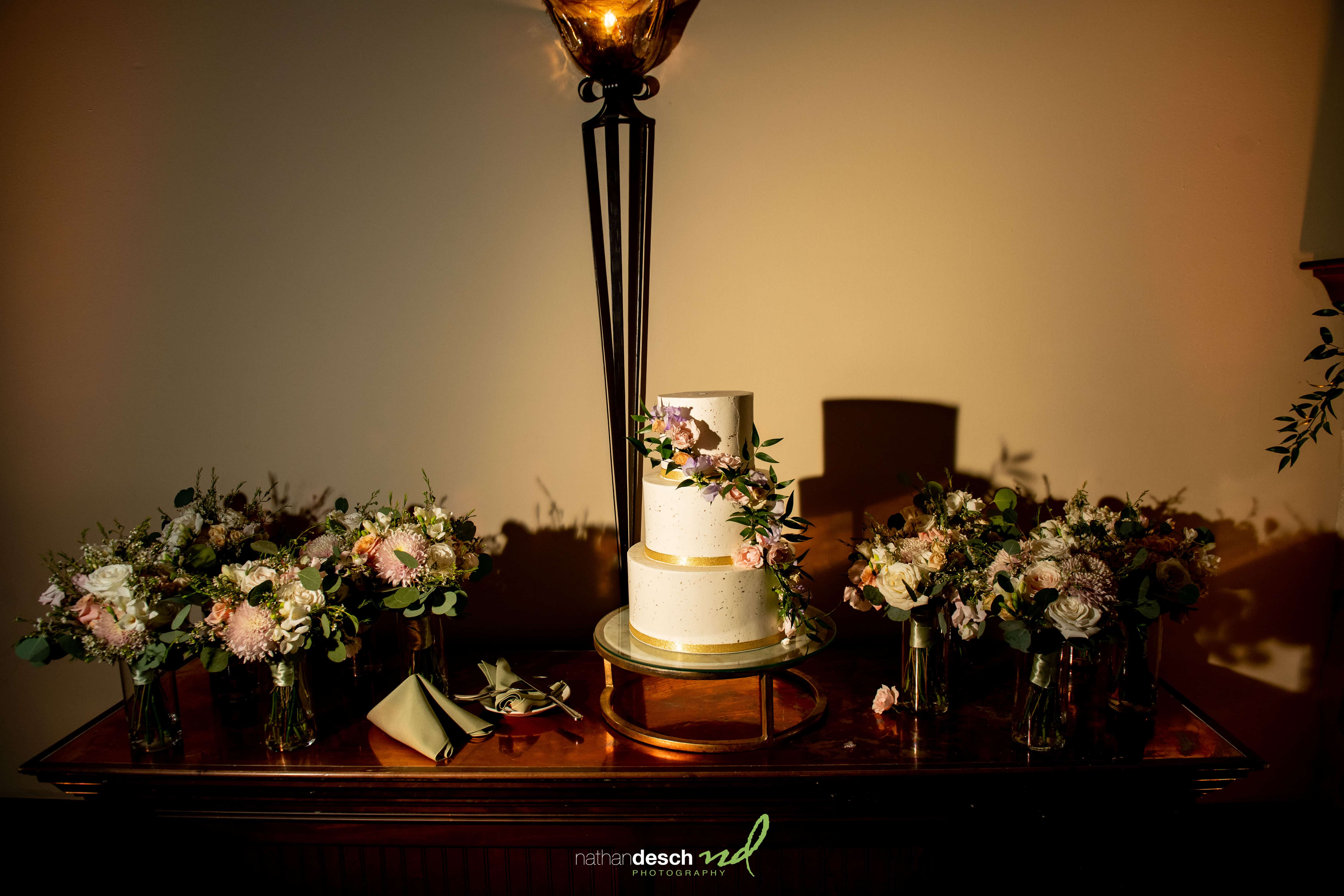 Two-tier white wedding cake with pink flowers on gold stand at The Inn at Leola Village, surrounded by spring bouquets.
