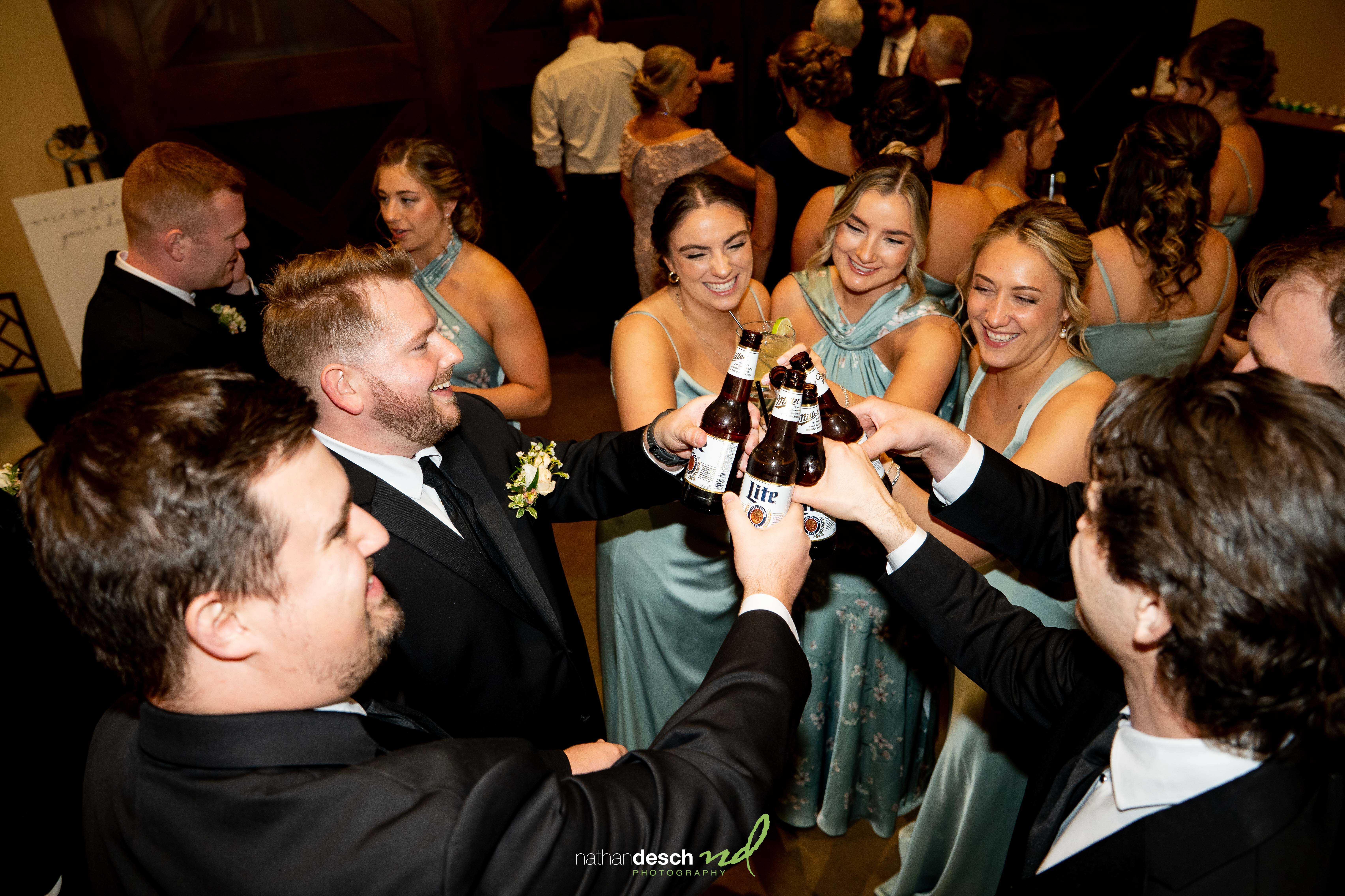 Wedding party in suits and teal dresses raise beer bottles for a joyful toast at the Inn at Leola Village reception.