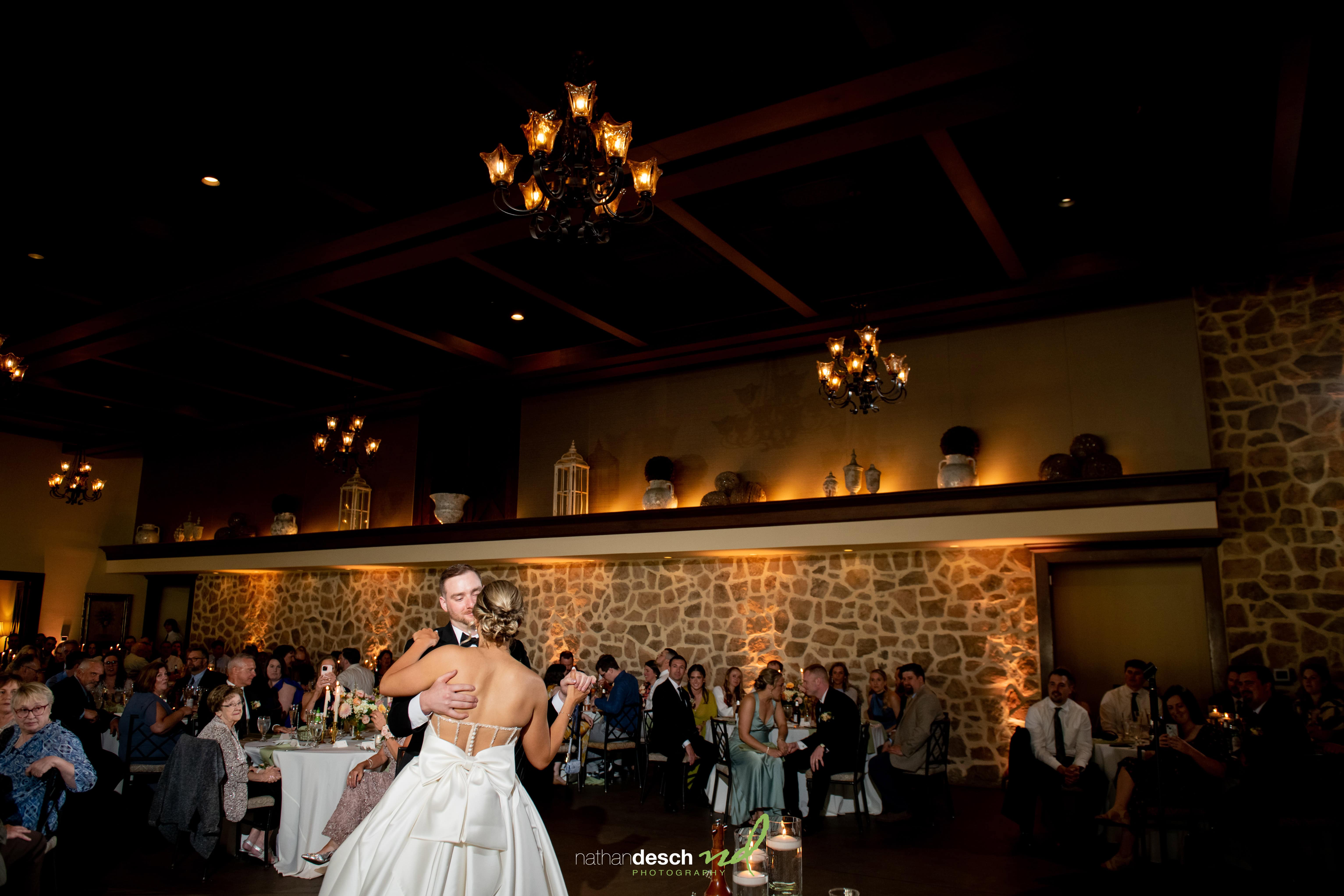 Bride and groom share first dance under chandeliers at the elegant Inn at Leola Village reception hall, guests watching joyfully.