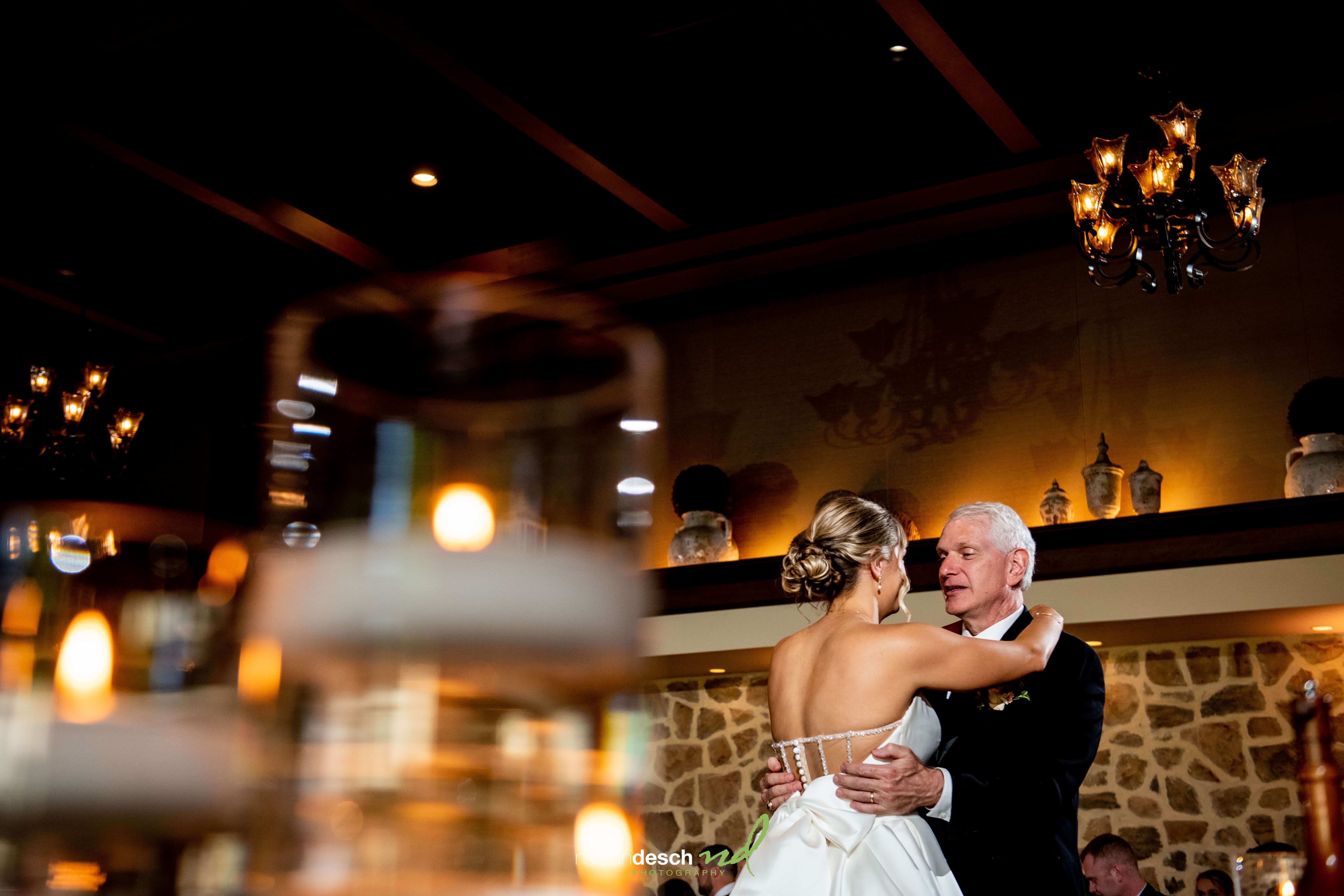 Bride dancing with her father at Inn at Leola Village, warm lighting, stone walls, chandeliers, romantic wedding atmosphere.