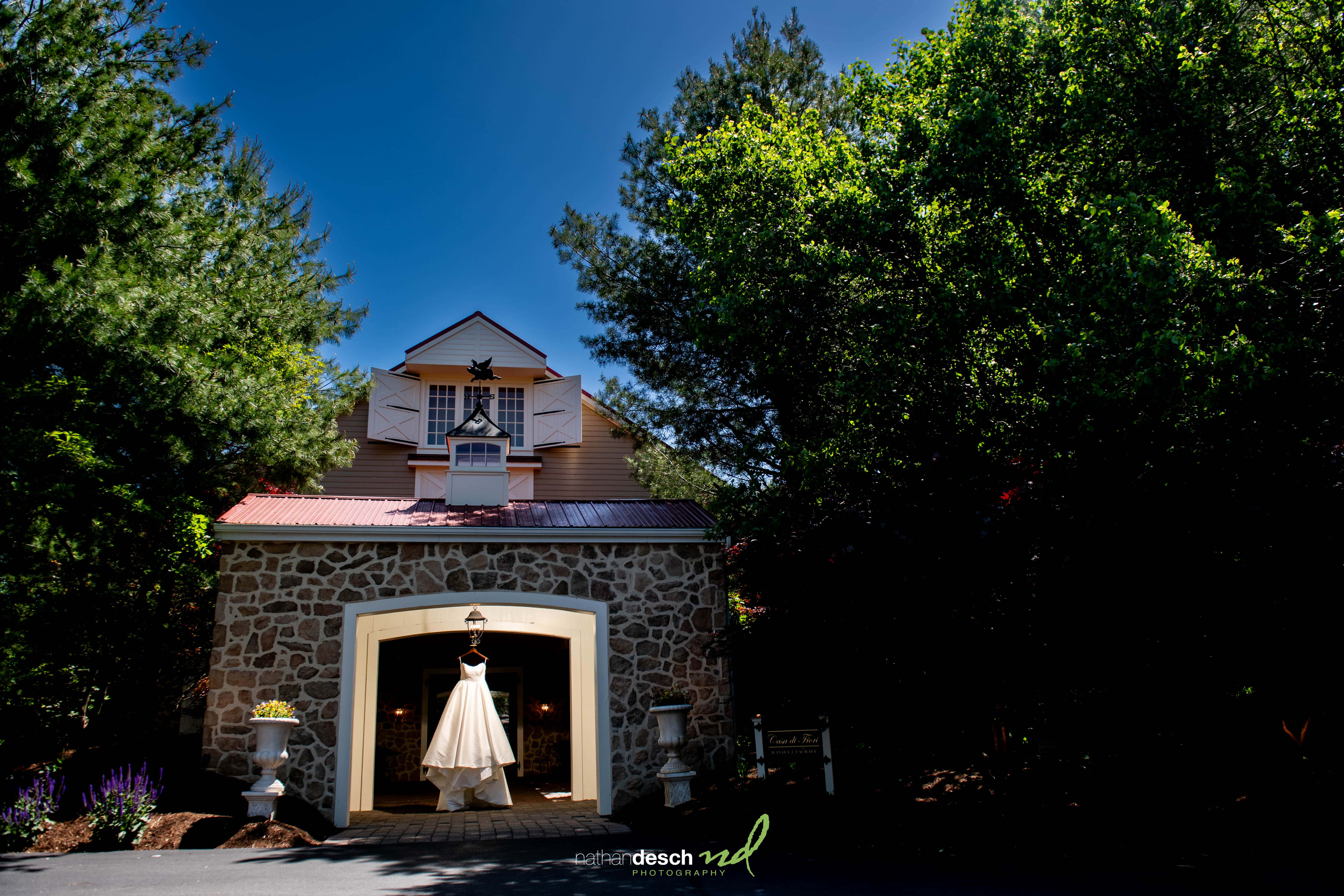 Elegant wedding dress hanging from lantern at rustic stone entrance, The Inn at Leola Village, with lush trees and blue sky.