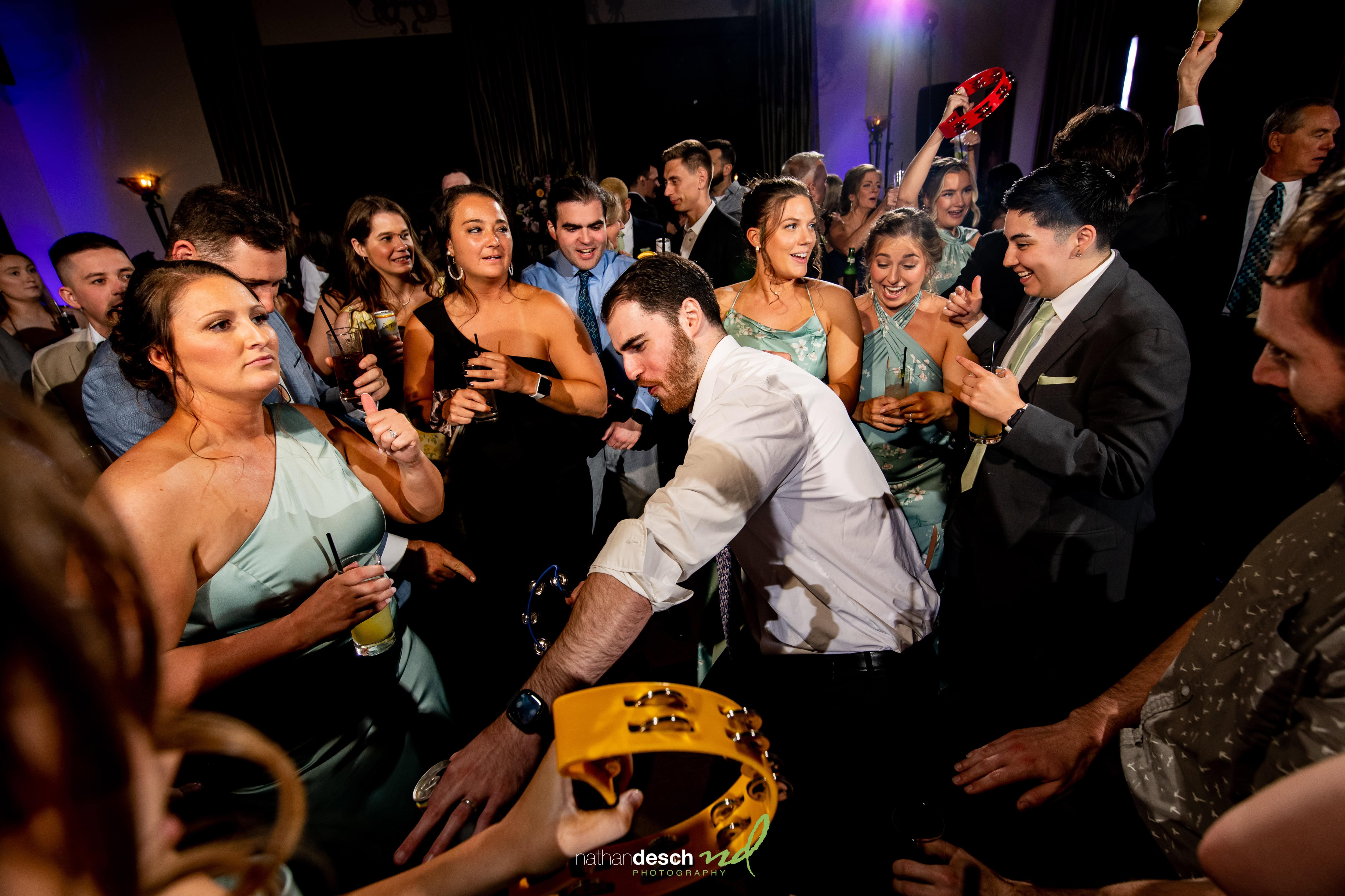 Wedding guests in formal attire joyfully dance with tambourines under colorful lights at the Inn at Leola Village reception.