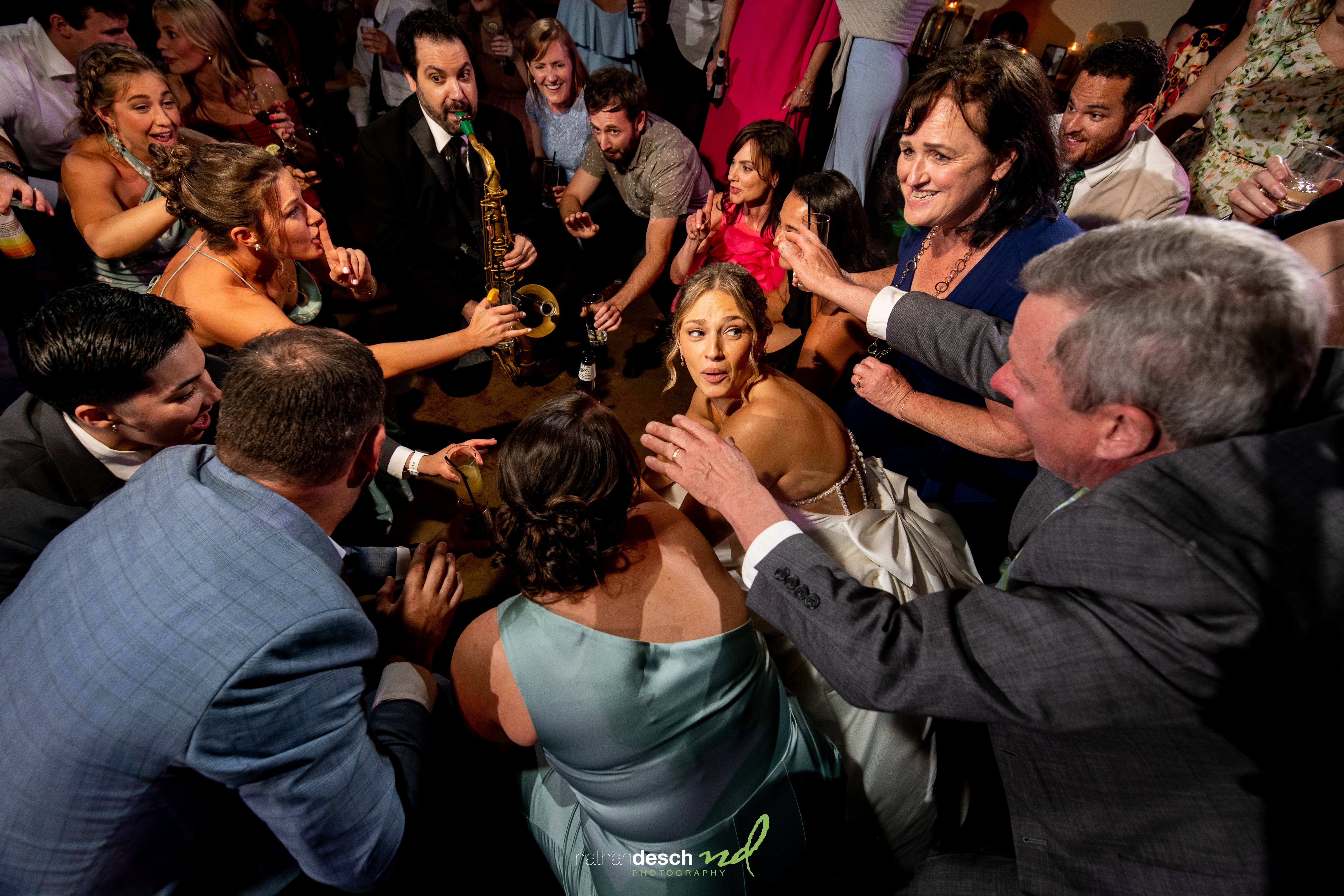 Bride in white dress surrounded by joyful wedding guests with live saxophone music.