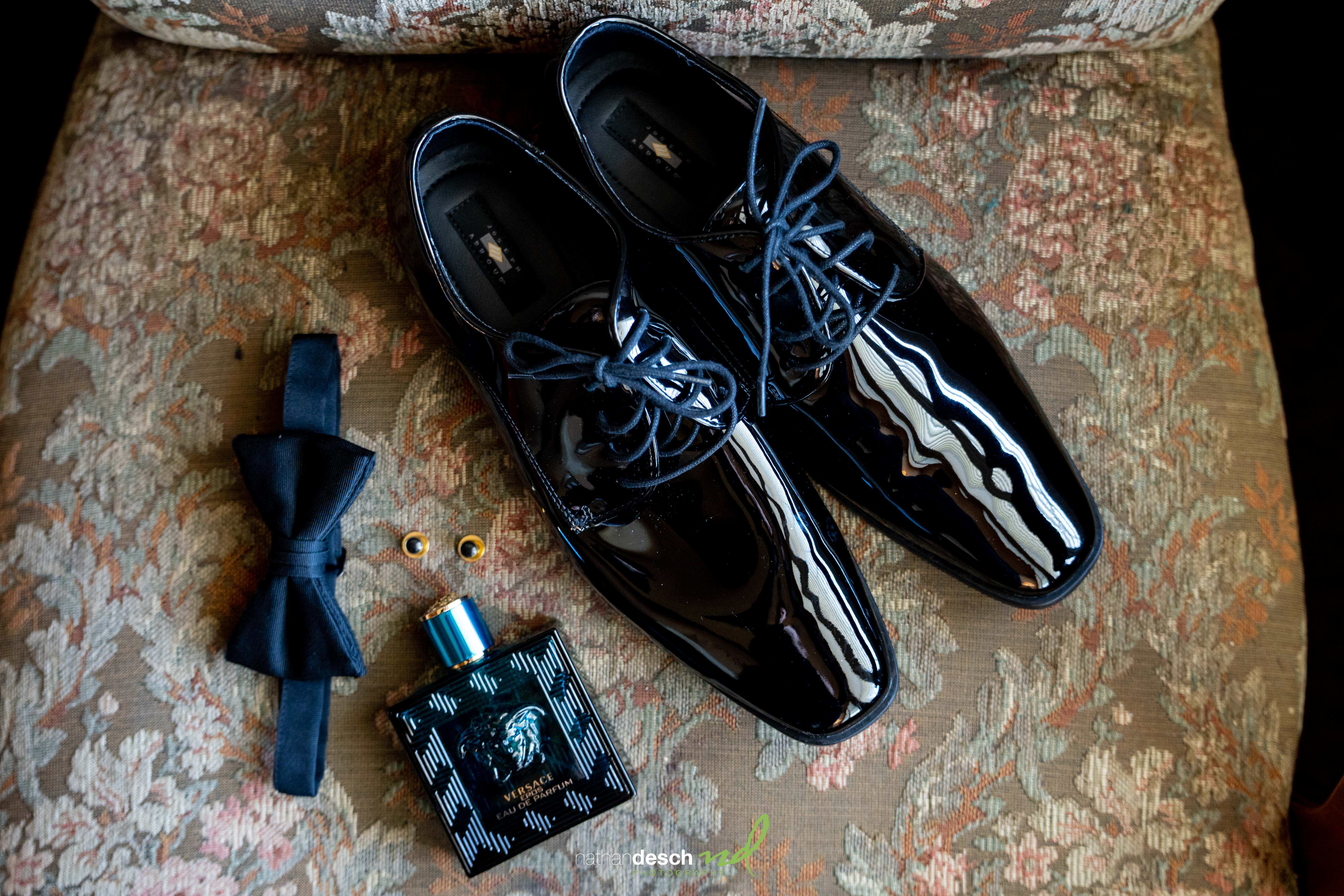Groom’s black dress shoes, bow tie, gold cufflinks, and Versace cologne arranged on chair at The Inn at Leola Village wedding.