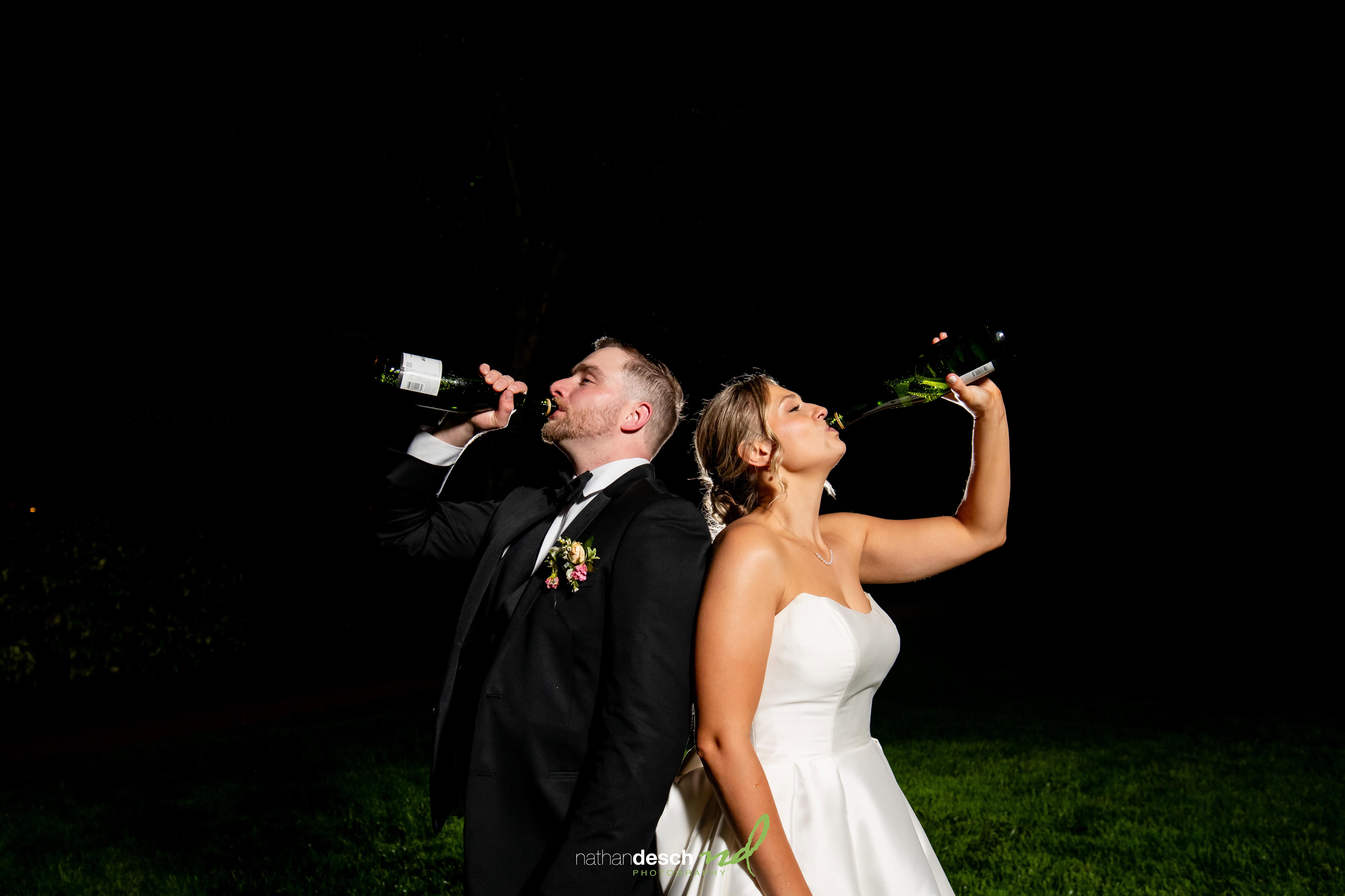 Bride and groom in elegant wedding attire laugh and drink wine back to back outdoors at night on grassy lawn.