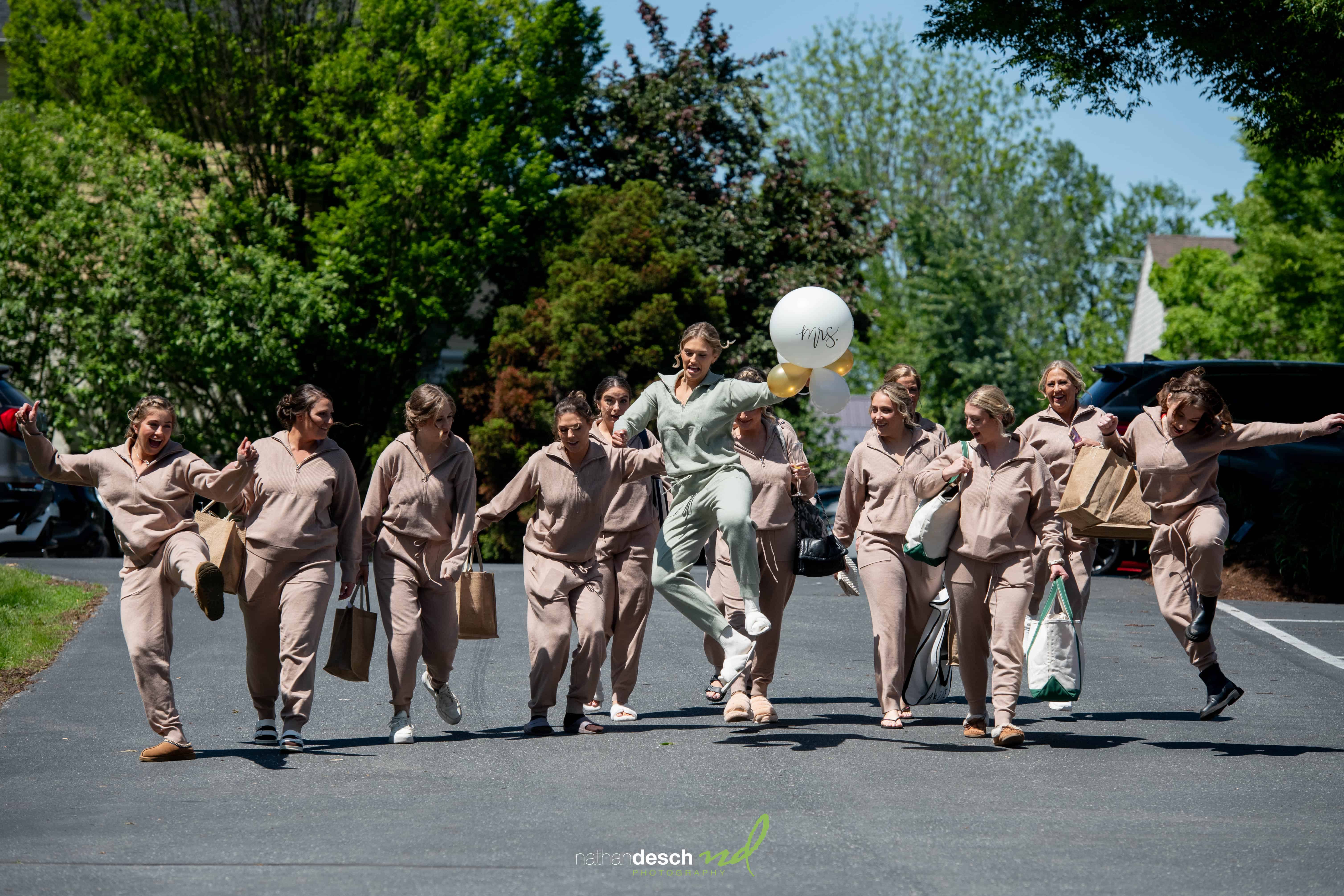 Bridesmaids in beige and bride in green jump joyfully with gift bags and Mrs. balloon on a sunny street at the Inn at Leola Village.
