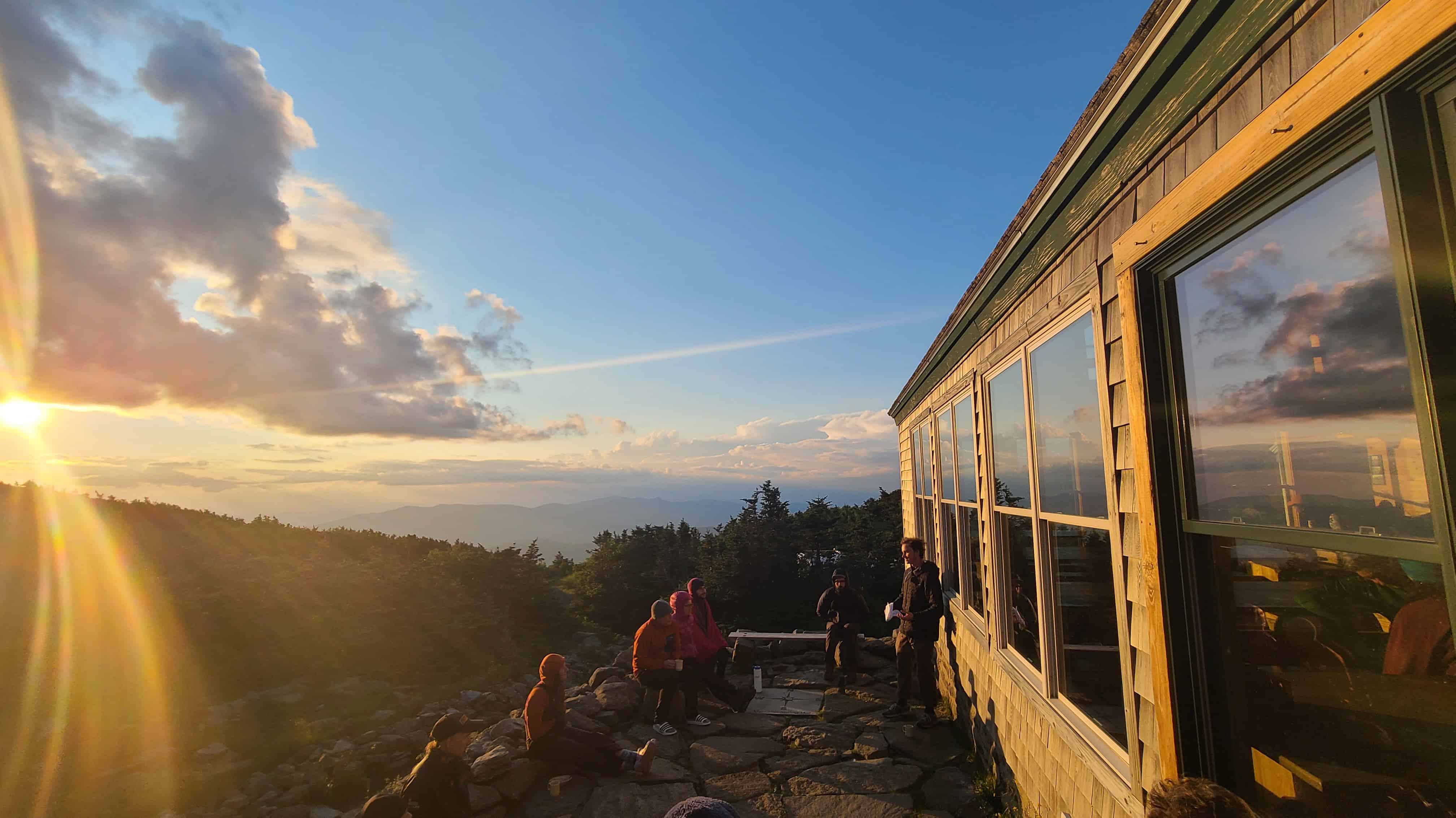 Adventure wedding party sits on rocks at sunset by wooden cabin, with mountain views and golden skies in the White Mountains.