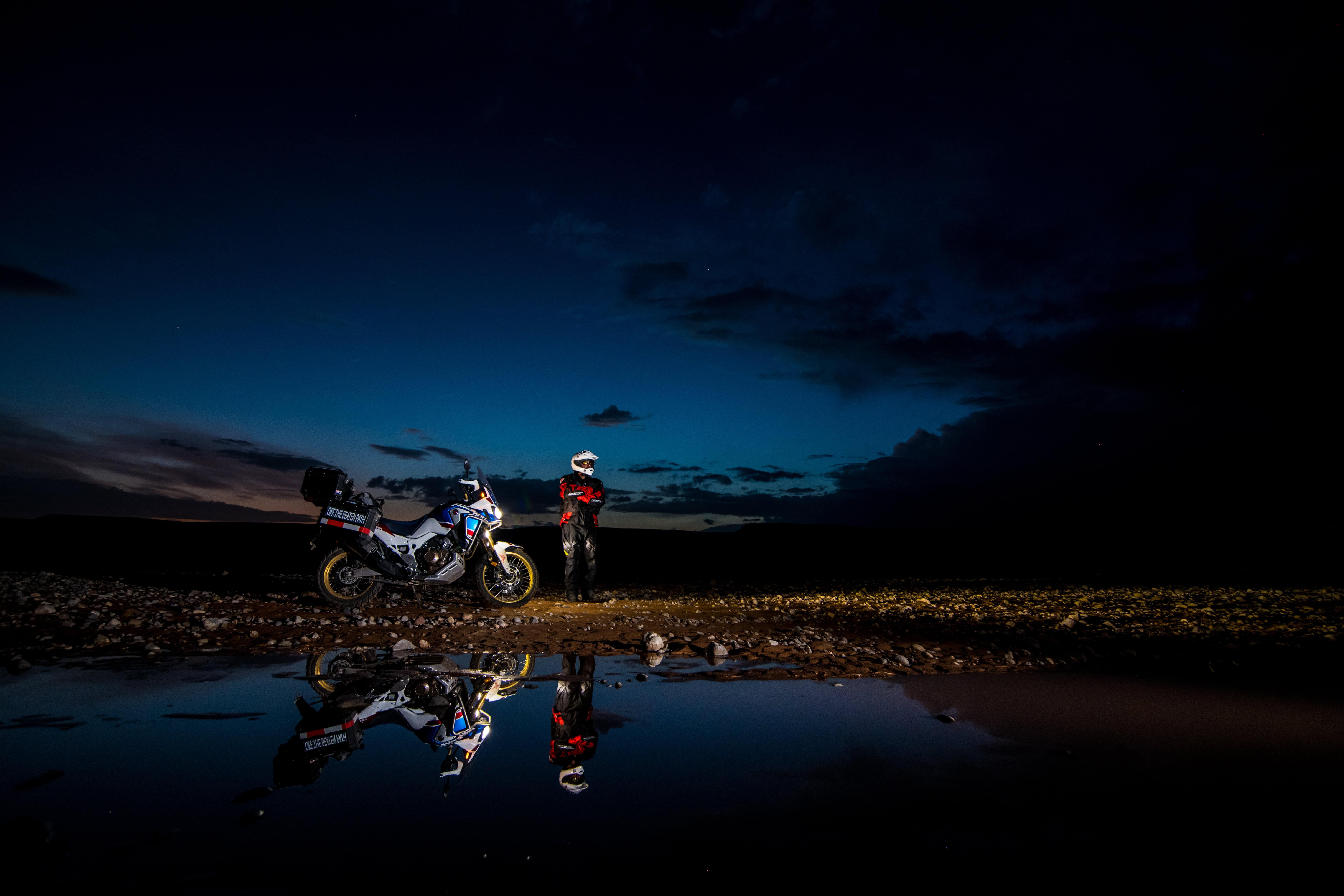 Bride and groom pose by a motorcycle at night in Marrakech, reflected in puddle against a romantic deep blue sky.