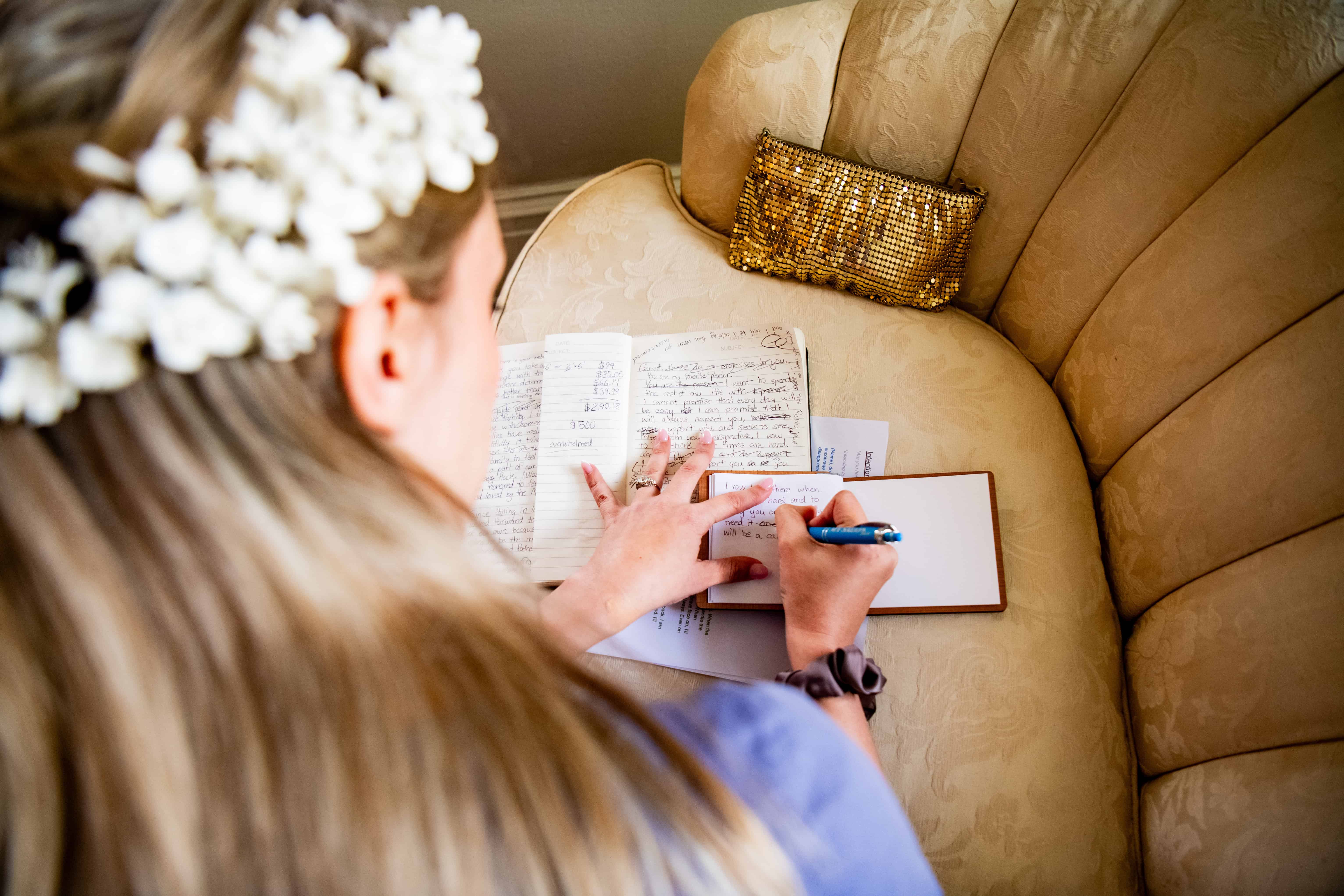 Bride with a floral hair accessory writes wedding photo tips in her notebook on a beige couch beside a gold sequined clutch.