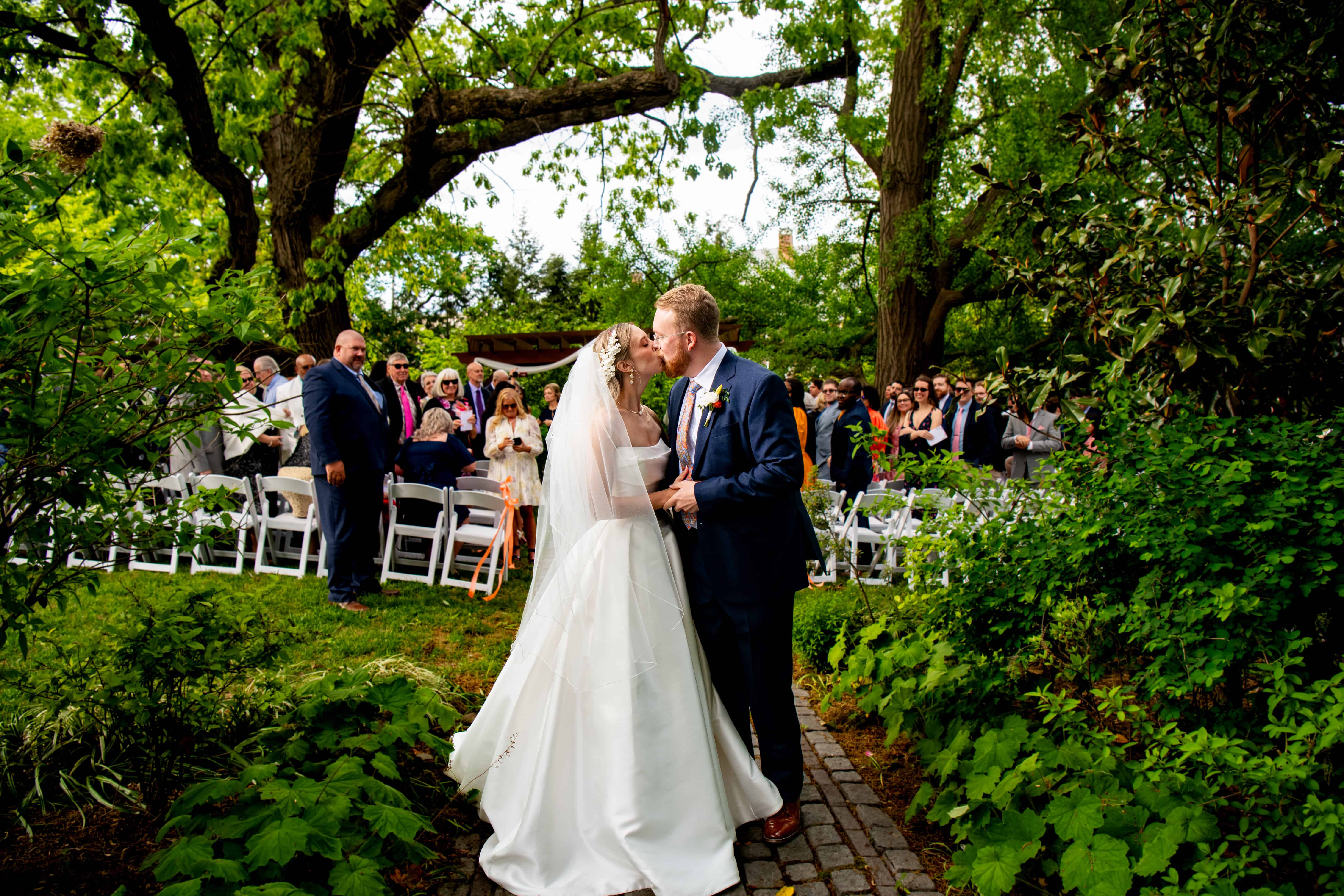 Bride and groom kiss on a lush garden path, joyful guests in formal attire watch outdoor wedding under tall trees.
