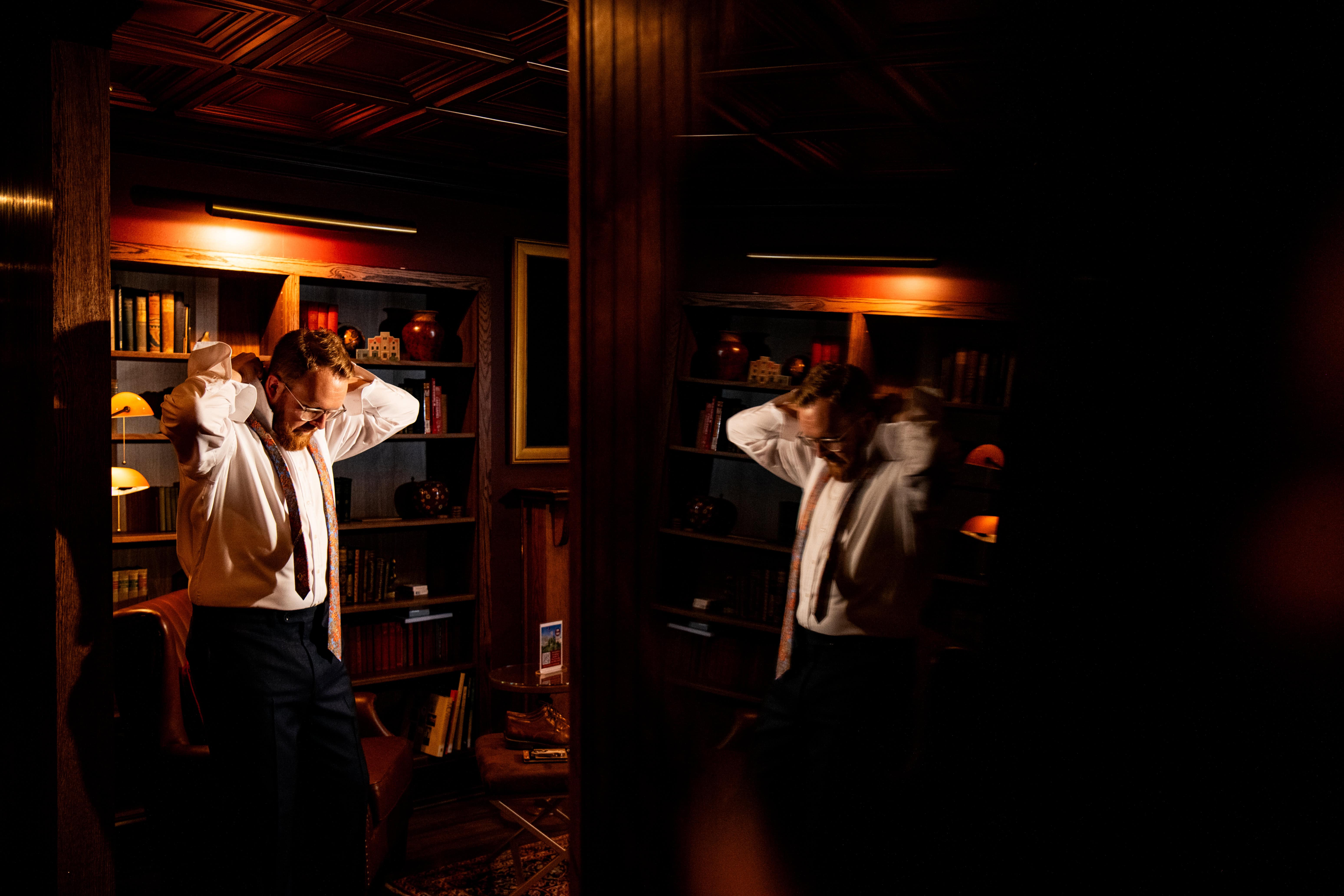 Groom in white shirt adjusts tie before wedding, standing by mirror in warmly lit room with bookshelves and wooden decor.