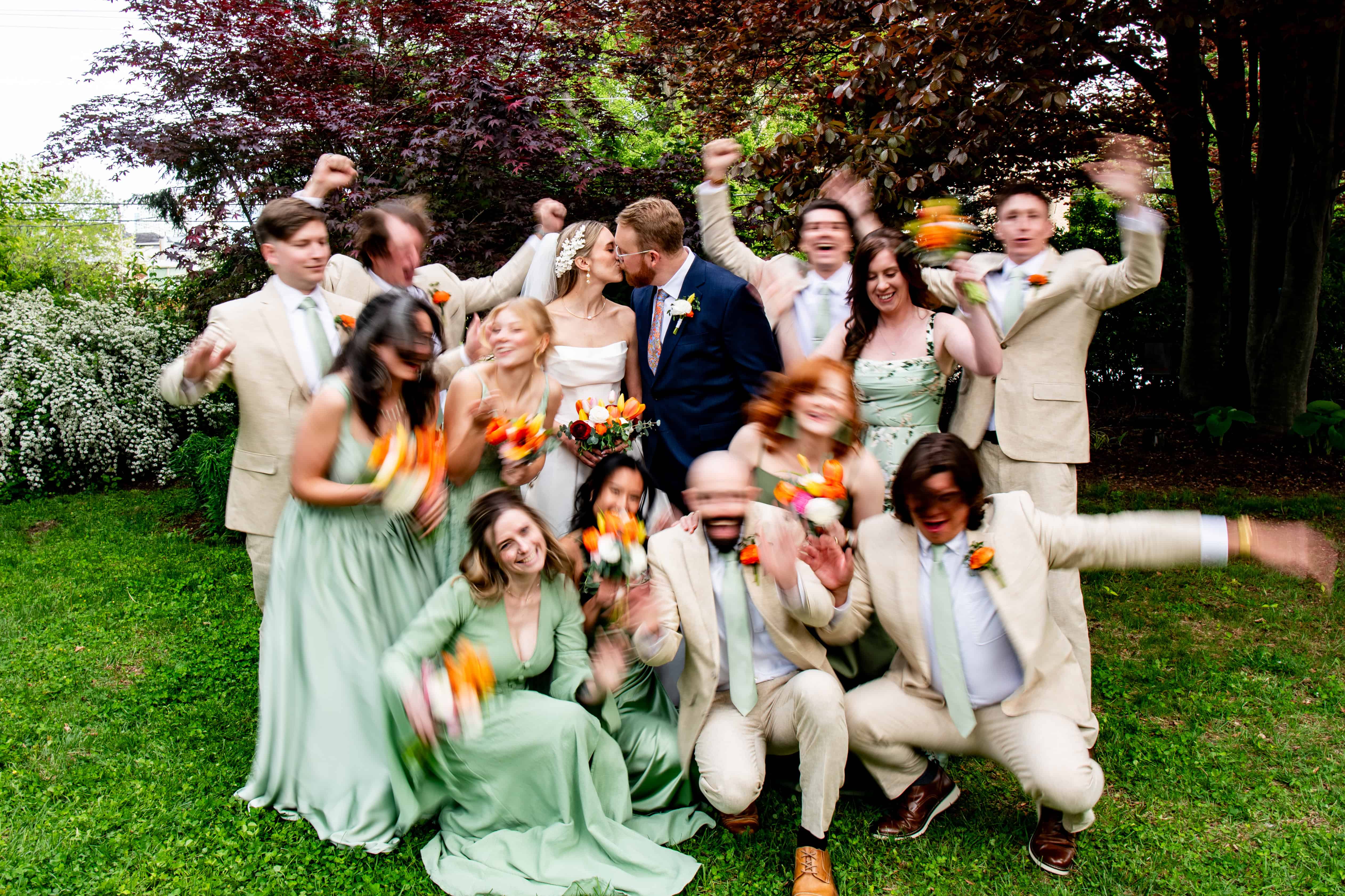 Bride and groom kiss in garden as wedding party in pale green dresses and tan suits cheer, natural wedding photography moment.